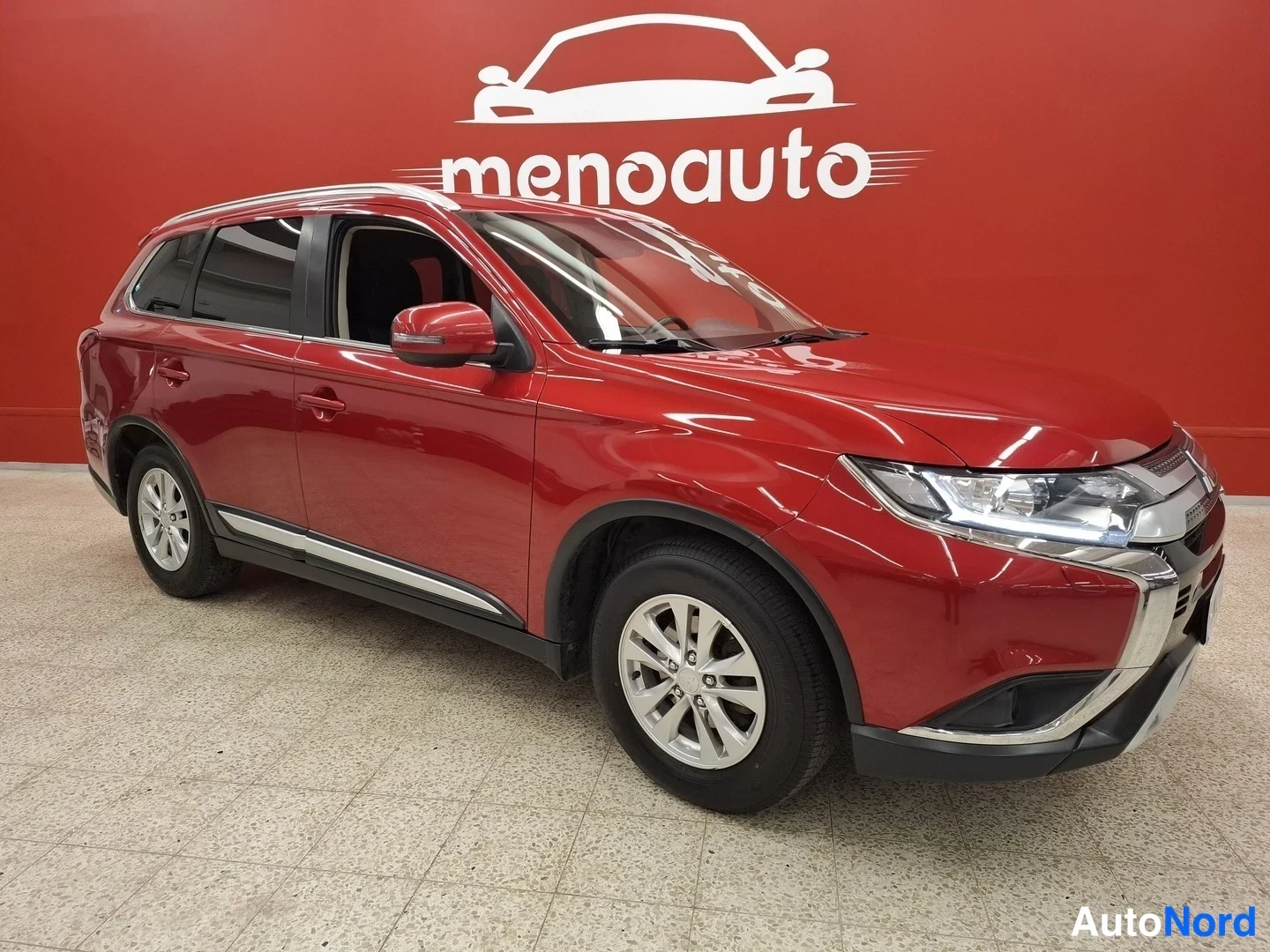 Etuviisto, nokka oikealle: Punainen Mitsubishi Outlander -maasturin etukulma sisätiloissa, keulagrami ja ajovalot näkyvät oikealla, sivut ja takapyörä vasemmalla.