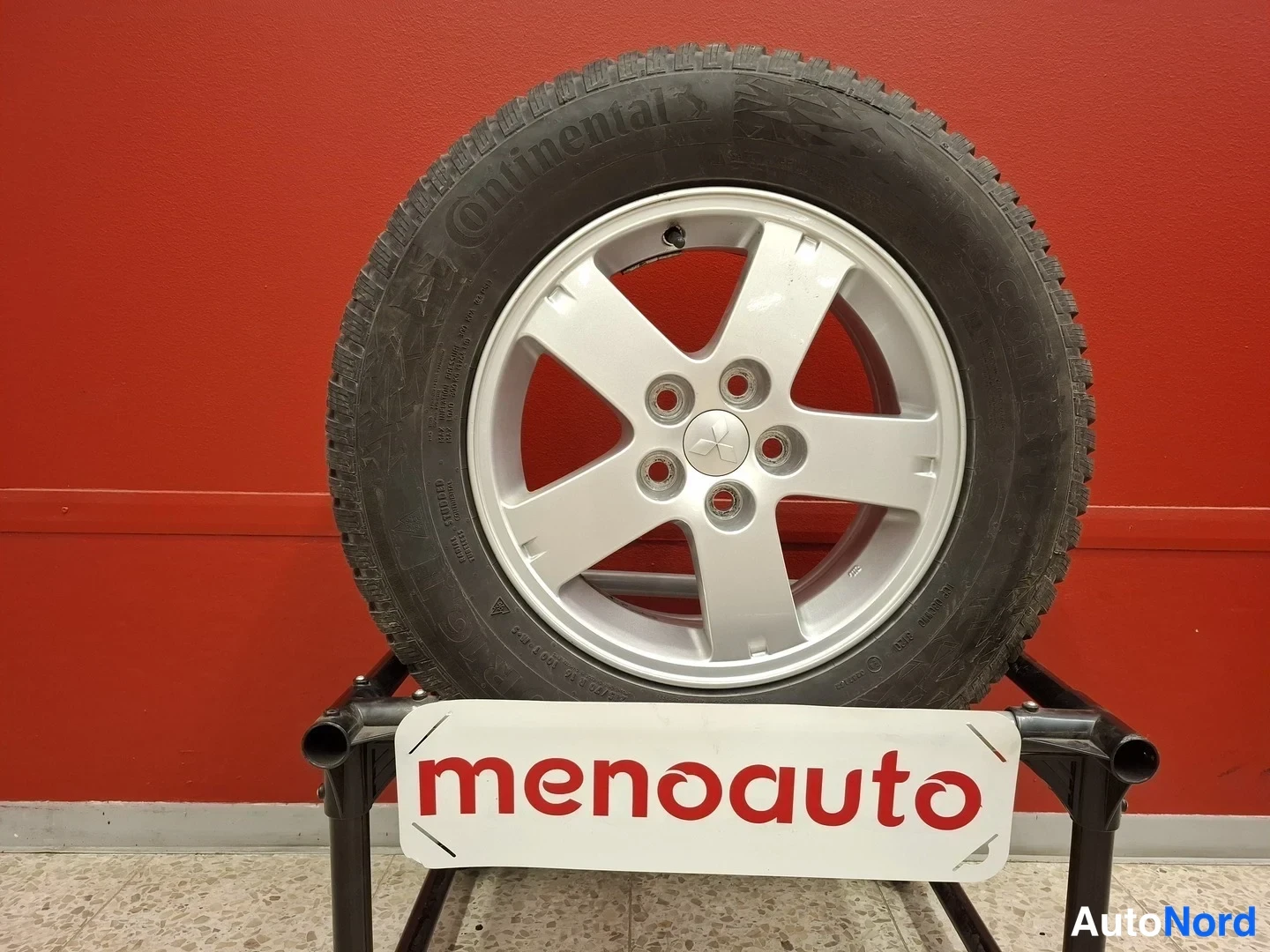 Sivulta: Continental-talvirenkaan studded tire on a silver alloy wheel on a stand, with a red 'menoauto' sign