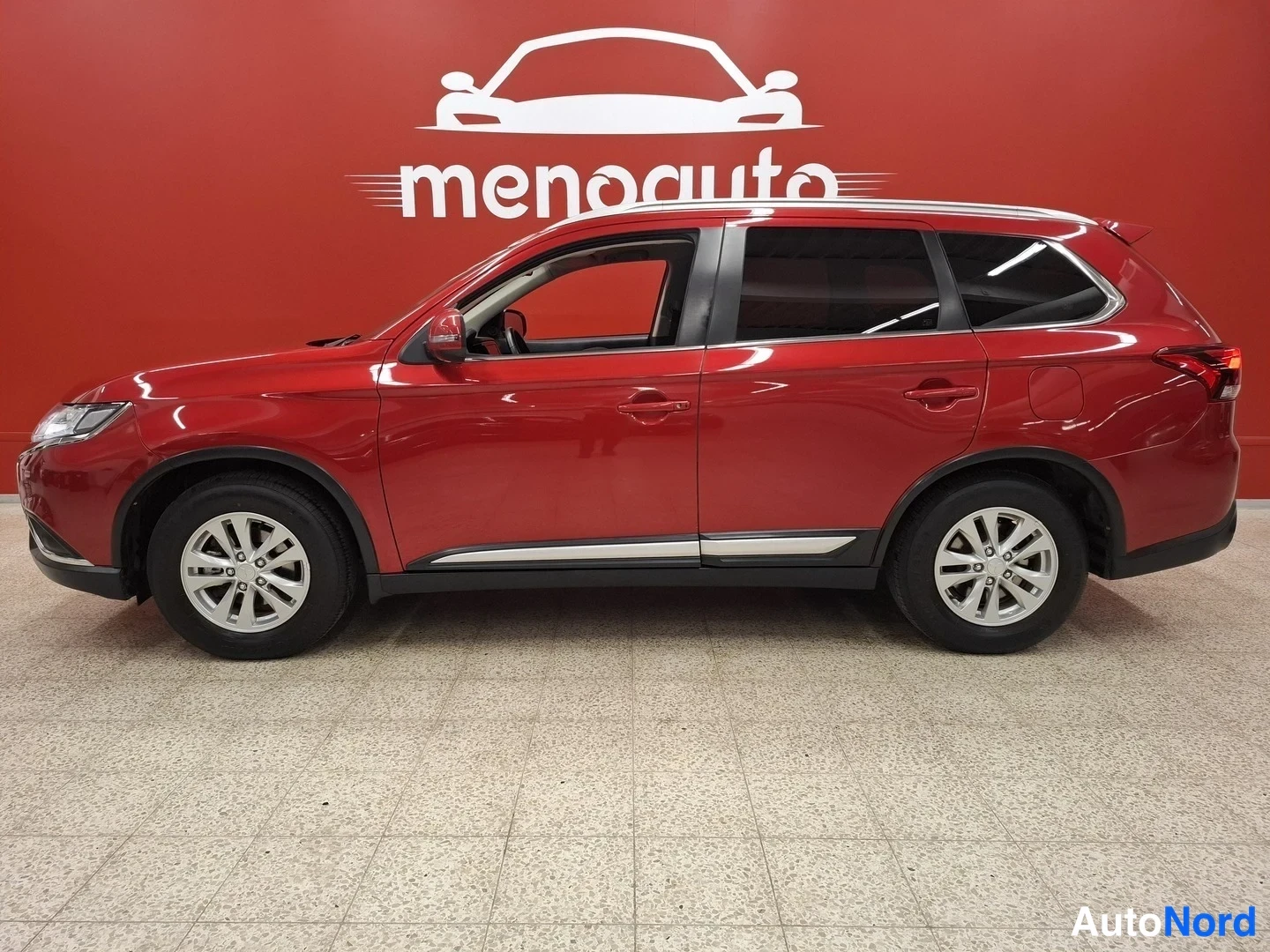 Etuviisto, nokka vasemmalle: Punainen Mitsubishi Outlander -maastokulkuvaltimo kuvattu edestä vasemmalla kolmasosakulmalla sisätiloissa punaista seinää vasten, kevytmetallivanteet