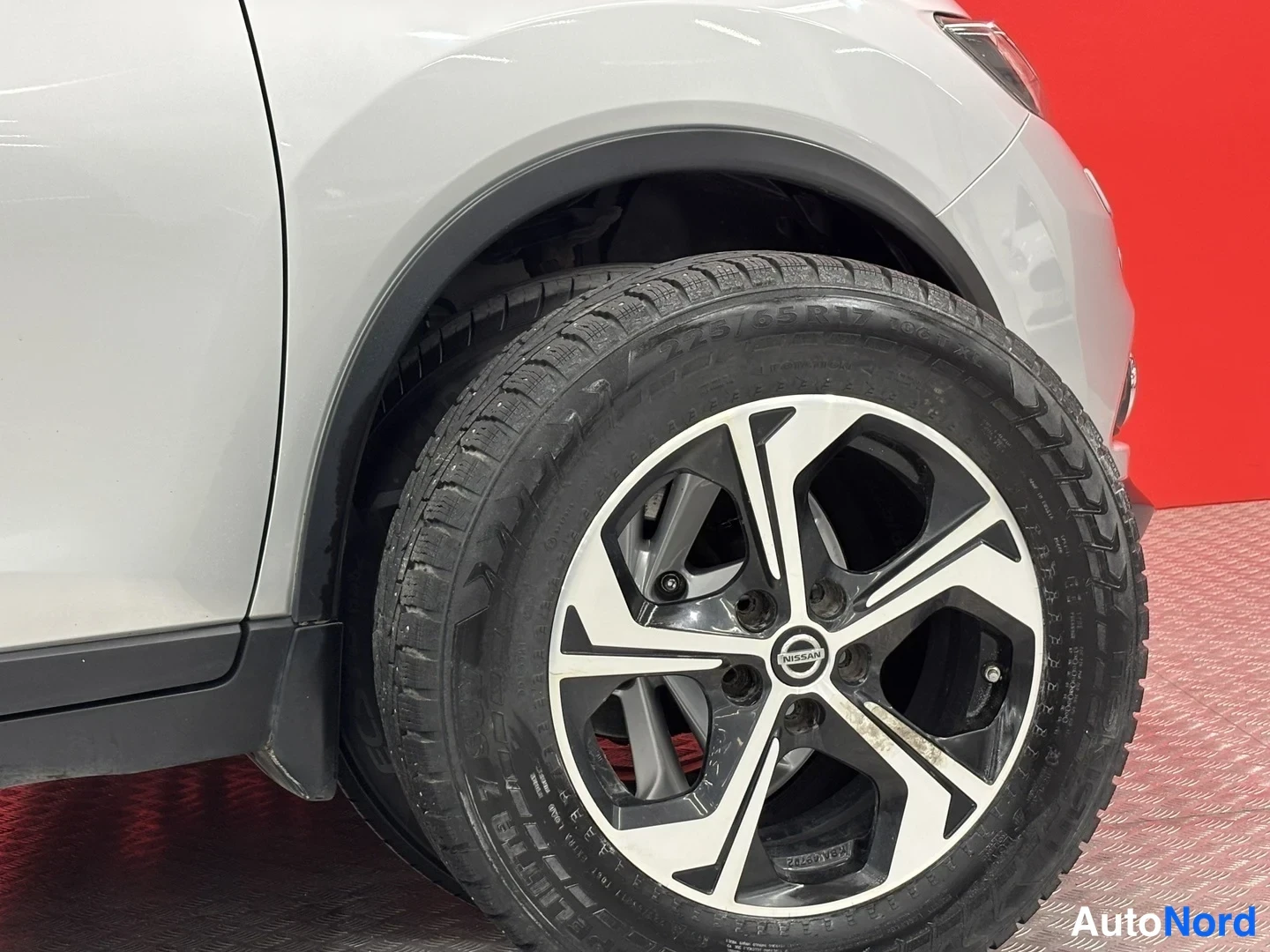 Etuviisto, nokka oikealle: Nissan-maasturin etupyörän lähikuva: hopeisen korin alle asennettu kattava mustavalkoinen alumiinivanne ja nastarenkaas, Nissan-merkki napissa.