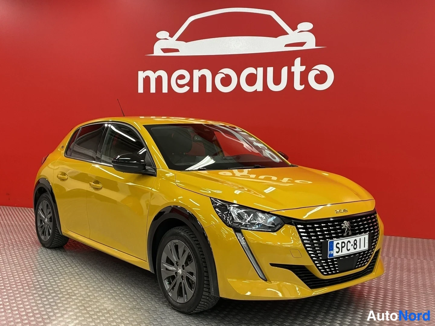 Etuviisto, nokka oikealle: Keltainen Peugeot 208 -henkilöauto kuvattu etukulmasta auton keulan ollessa kuvan oikeassa laidassa, punaisella menoauto-mainosseinämä taustalla.