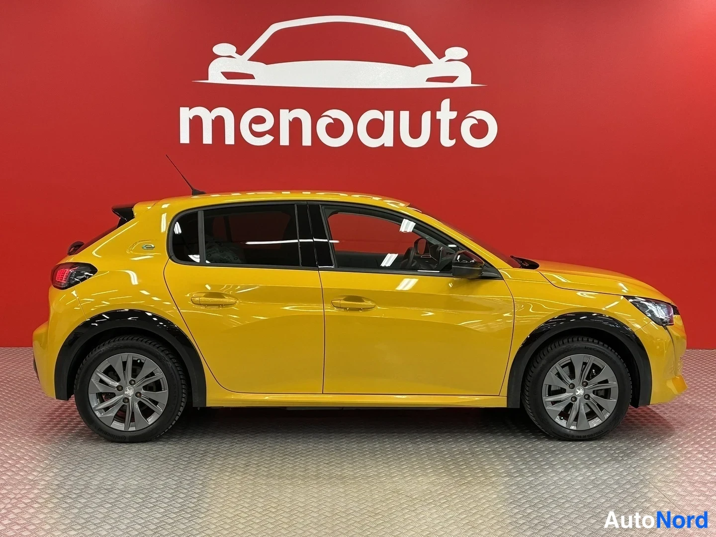 Etuviisto, nokka oikealle: Keltainen Peugeot 208 -henkilöauto sivukulmasta keulan suunnattuna oikealle, punainen menoauto-mainosseinämä taustalla.