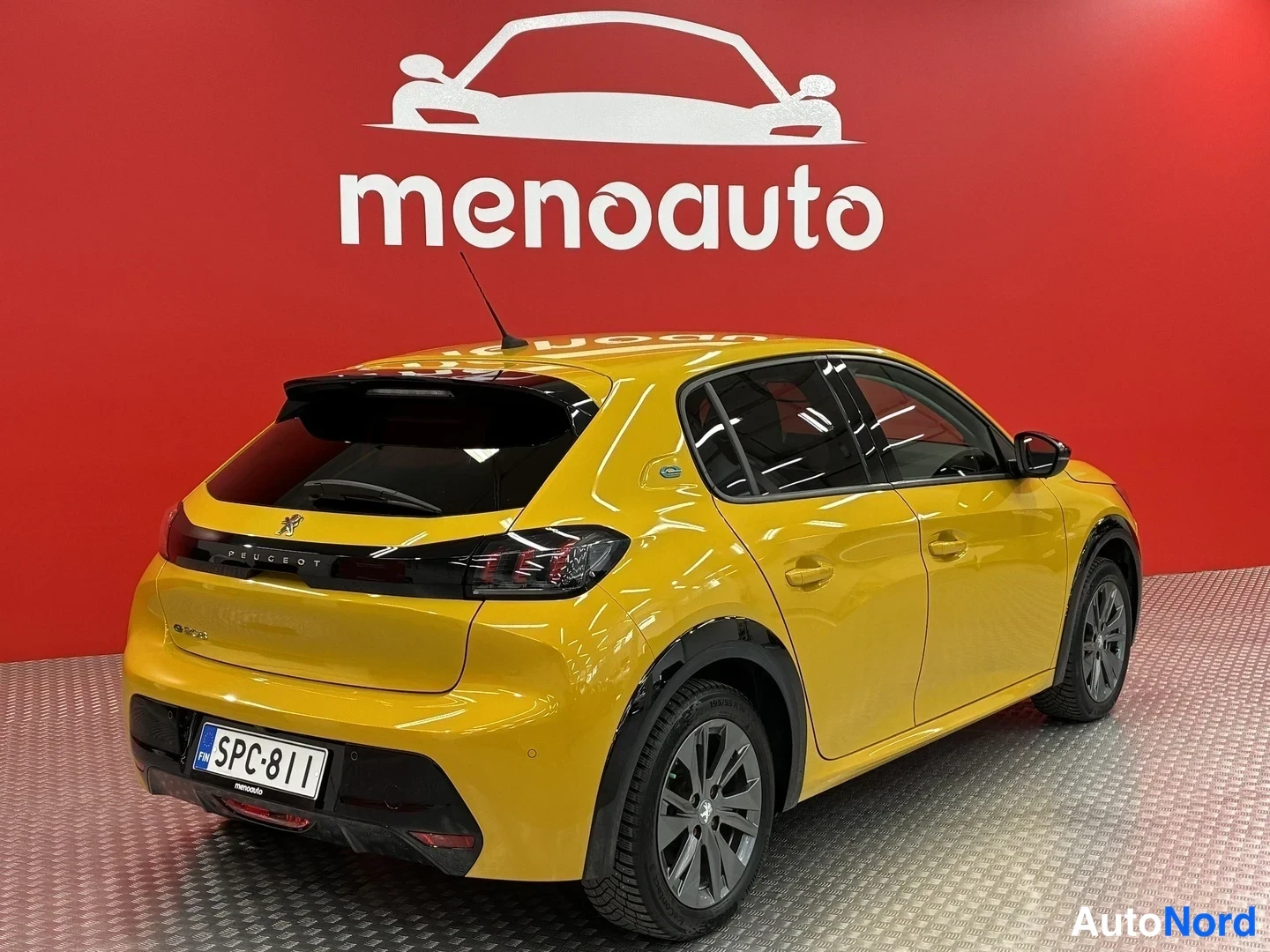Takaviisto: Keltainen Peugeot 208 -henkilöauto takakulmasta, rekisterikilpi, punainen menoauto-mainosseinämä taustalla.