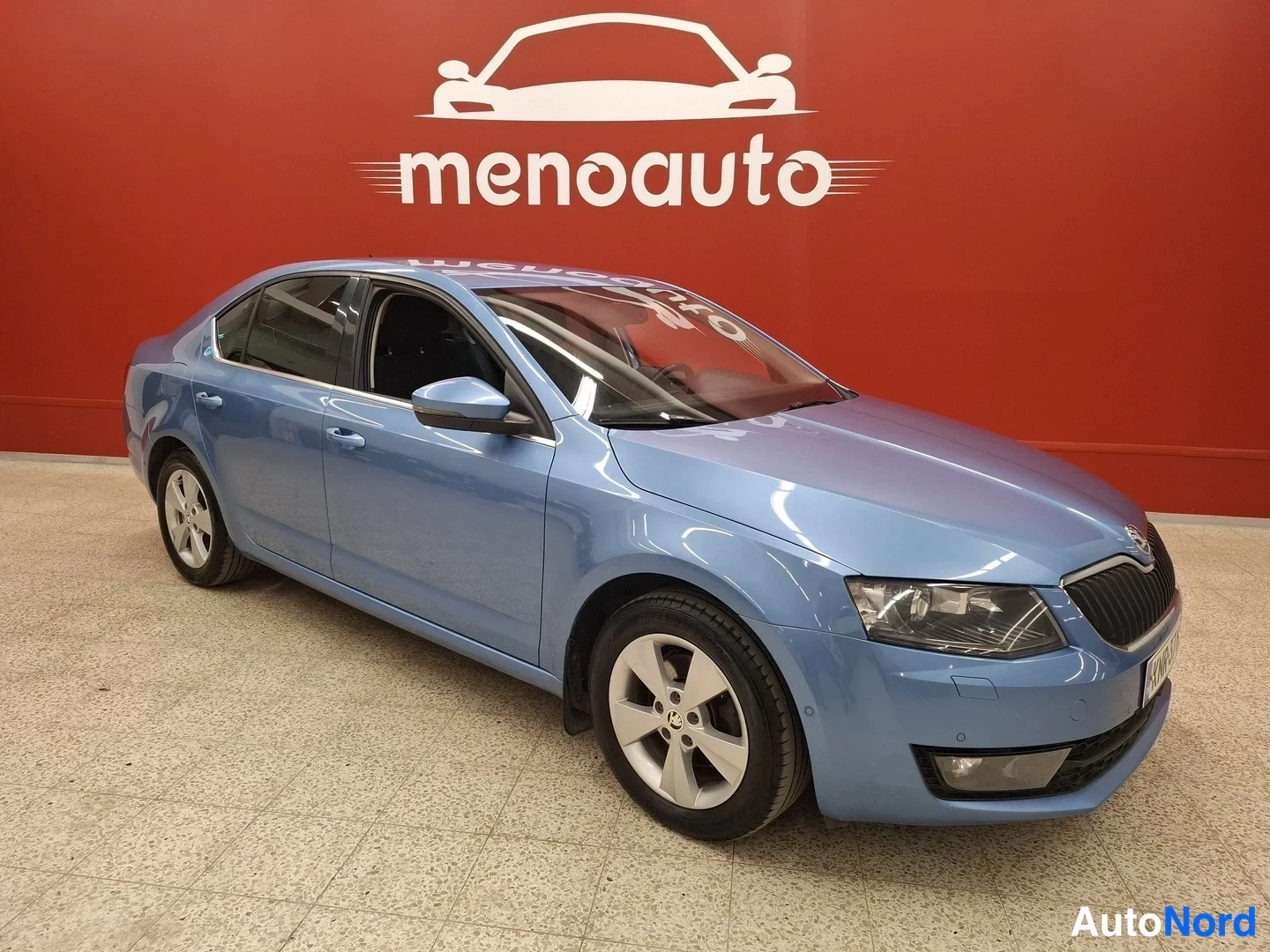 Etuviisto, nokka oikealle: Sininen Skoda Octavia -henkilöauto kuvattu kolmannelta etukulmalta, keula oikealla, sisätiloissa punaisen seinän ja laatoitetun lattian edessä.