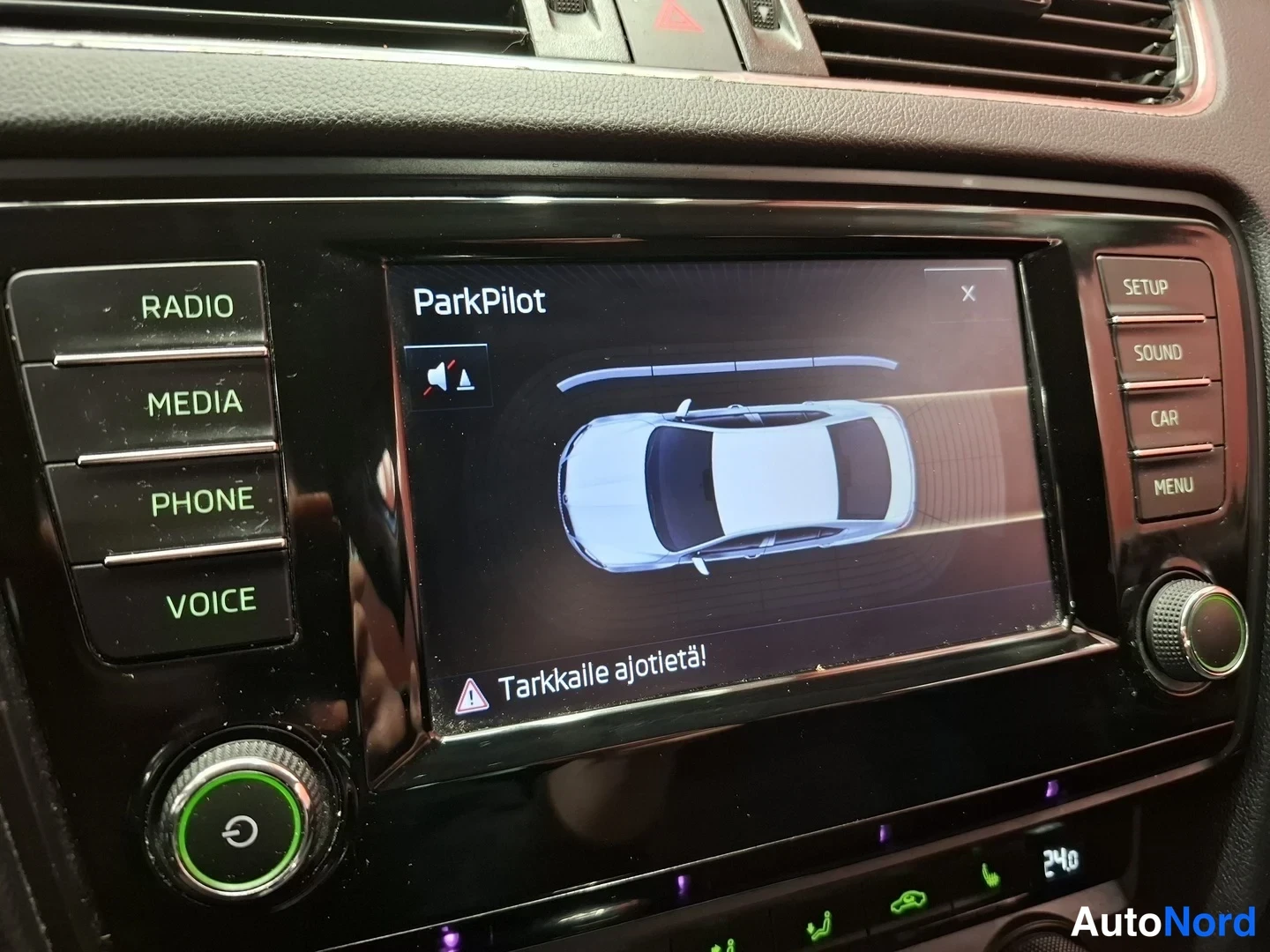 Muu: Kojelaudan kosketusnäyttö näyttää ParkPilot-törmäysvaroitusviestin ja auton yläkuvan, ympärillä radio- ja media-painikkeet.