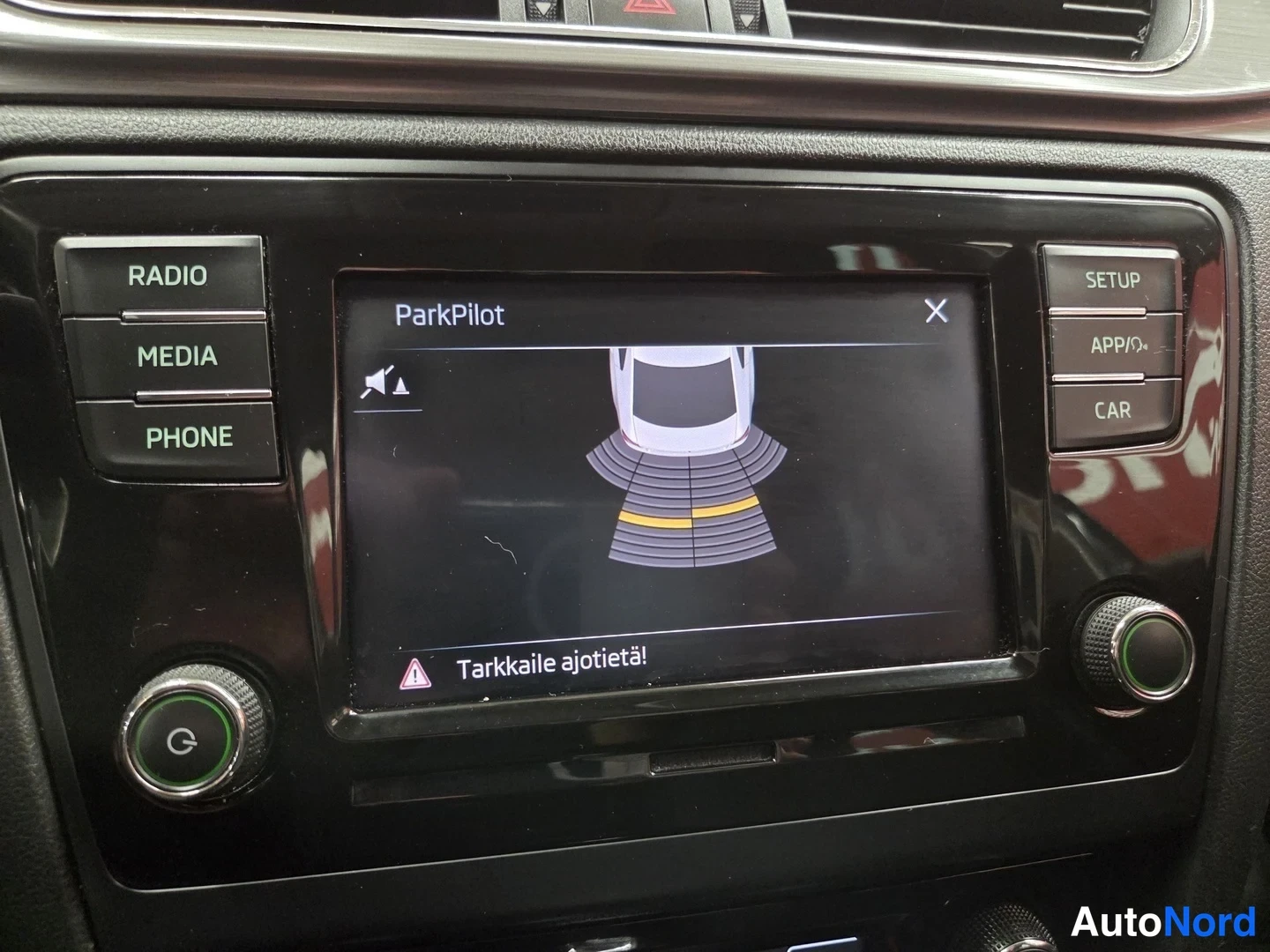 Yksityiskohta: Infotainment-näyttö ParkPilot-törmäysvaroitustoiminnossa: auton kuva ja anturikaaret, alareunassa varoitus 'Tarkkaile ajotietä!'.
