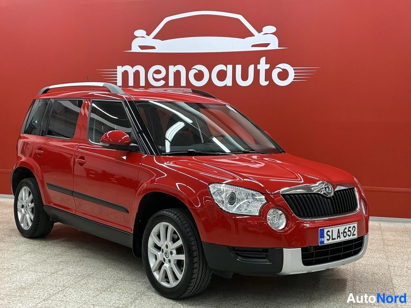 Etuviisto, nokka oikealle: Punainen Skoda Yeti etukulmasta oikealta, rekisteritunnus, taustalla punainen menoauto-mainosseina.
