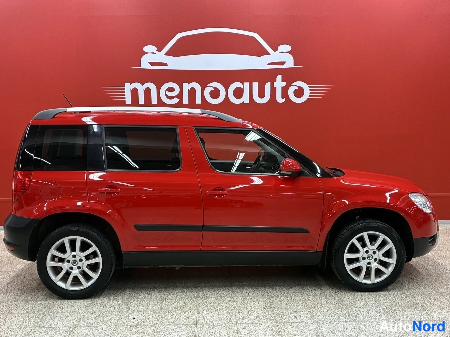 Sivulta: Punainen Skoda Yeti sivulta vasemmalta, hopeavanteet, taustalla punainen menoauto-mainosseina.