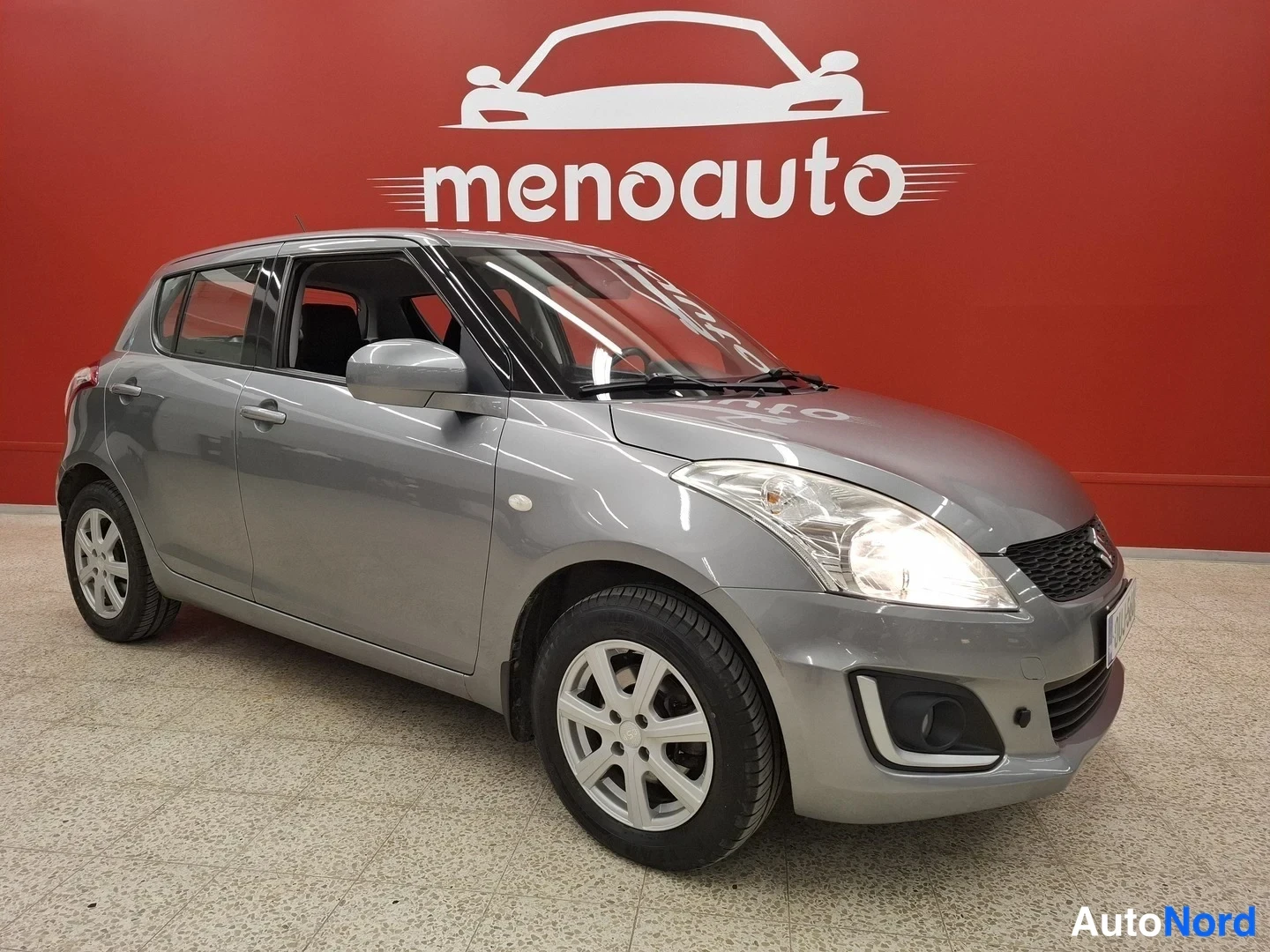 Etuviisto, nokka oikealle: Harmaa viistosti edestä oikealta kuvattu Suzuki Swift -henkilöauto sisätiloissa.