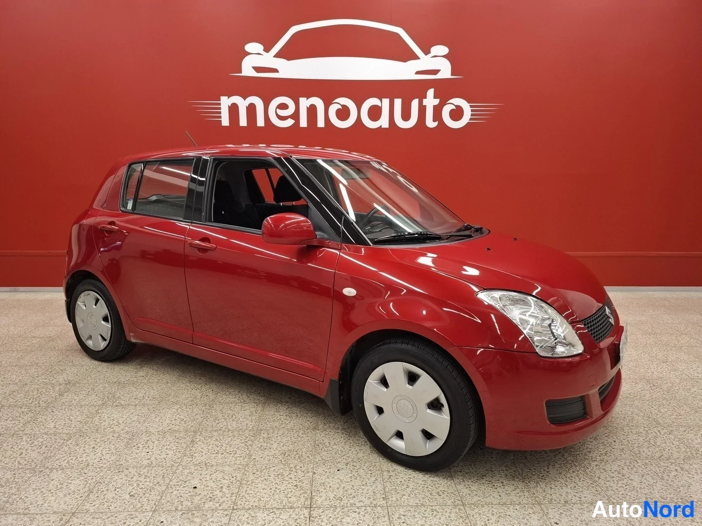 Etuviisto, nokka oikealle: Punainen viisiovinen Suzuki Swift etuviistossa oikealta, hopeiset peltivanteet, punainen studioseinä menoauto-logolla taustalla.