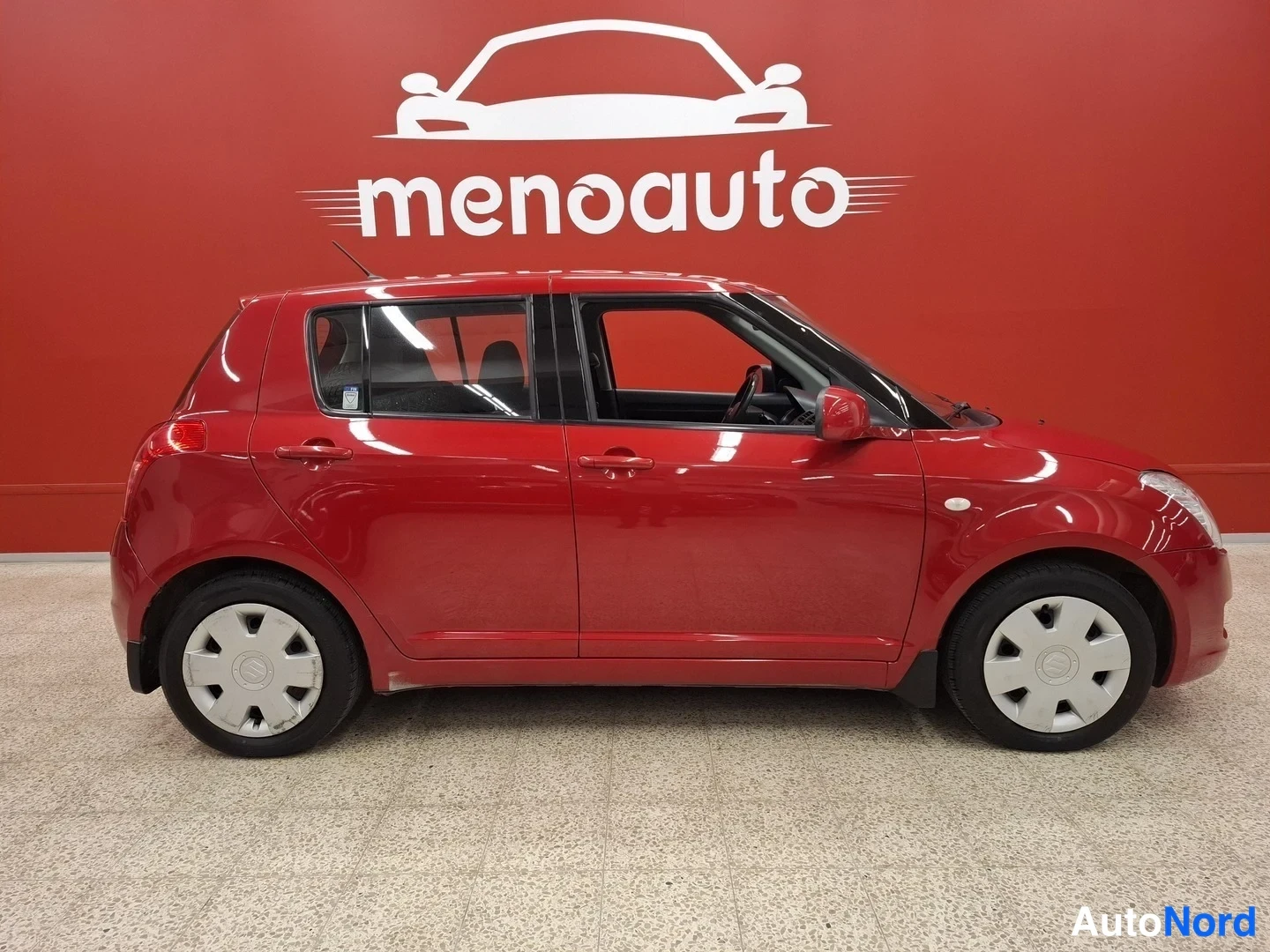 Sivulta: Punaisen Suzuki Swiftin sivuprofiili, oikeanpuoleiset ovet ja pyörät näkyvät punaista studioseinaä vasten.