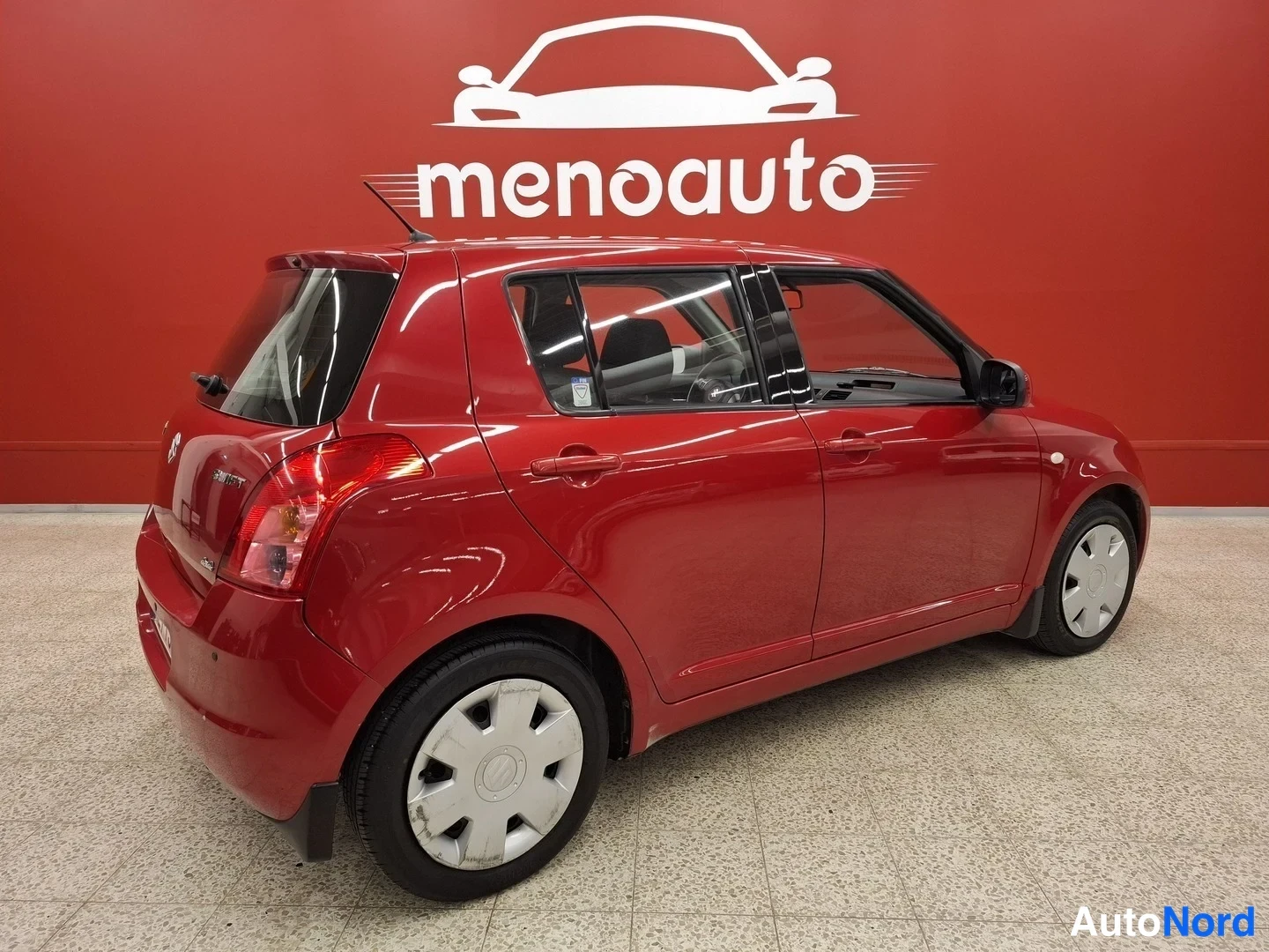 Takaviisto: Punainen Suzuki Swift takaviistossa oikealta, takavalot ja Takimmainen ovi näkyvät punaisen seinän edessä.