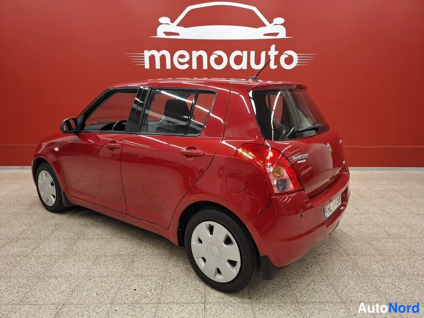 Takaviisto: Punaisen Suzuki Swiftin takakolmasosa sisätiloissa, perä ja takavalot näkyvissä, rekisterikilpi kyltinmyyjän baanassa.
