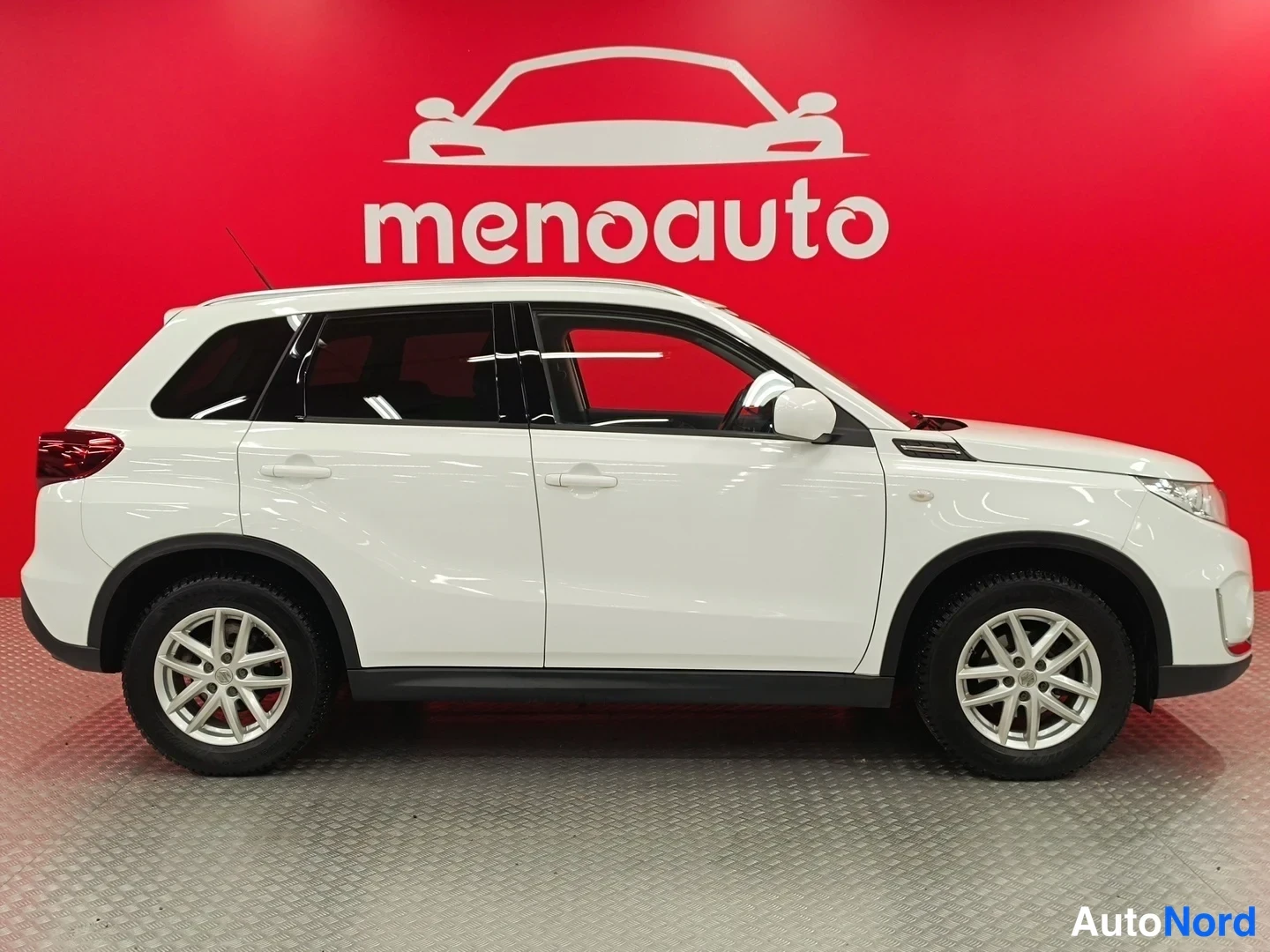 Etuviisto, nokka oikealle: Valkoinen Suzuki Vitara -maastauto sivukulmasta vasemmalta, menoauto-taustaa vasten.