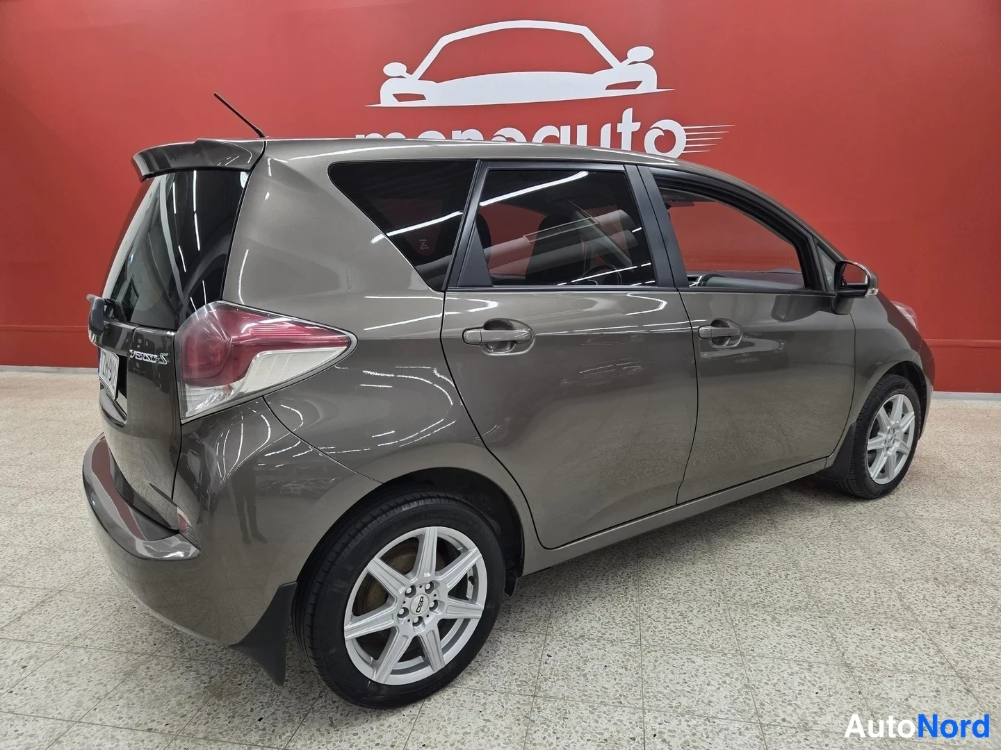 Takaviisto: Toyota Verso-S -henkilöauto kolmestinäkymästä takaa vasemmalta, harmaanruskea, sisätiloissa punaisella menoauto-taustalla.
