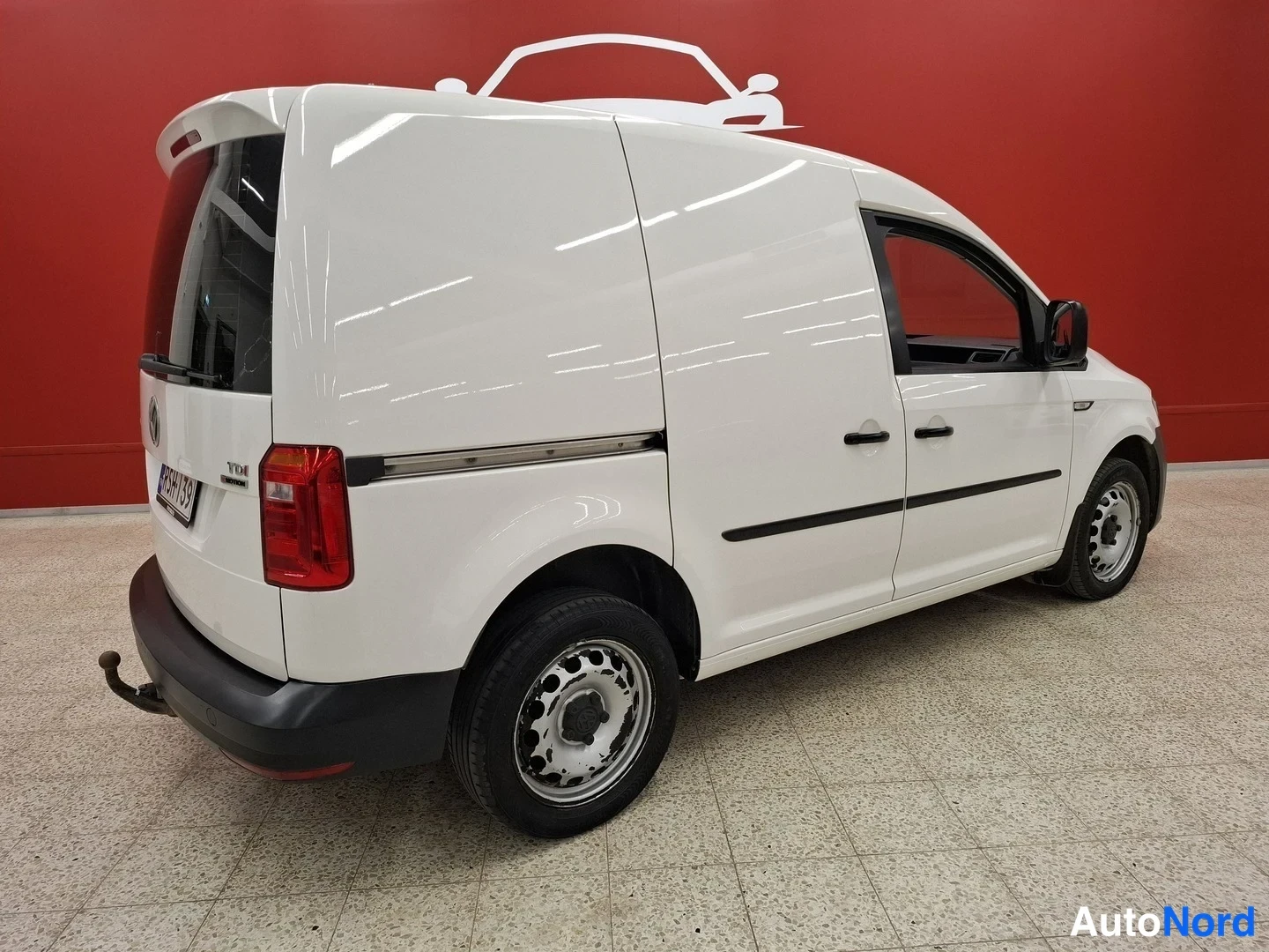 Takaviisto: Valkoinen pakettiauto takakulmasta, perä vasemmalla, punainen taustaseinä.