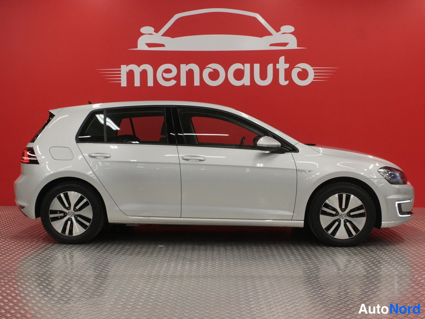 Sivulta: Valkoinen Volkswagen Golf sivulta vasemmalta, punainen taustaseinä ja menoauto-logo.