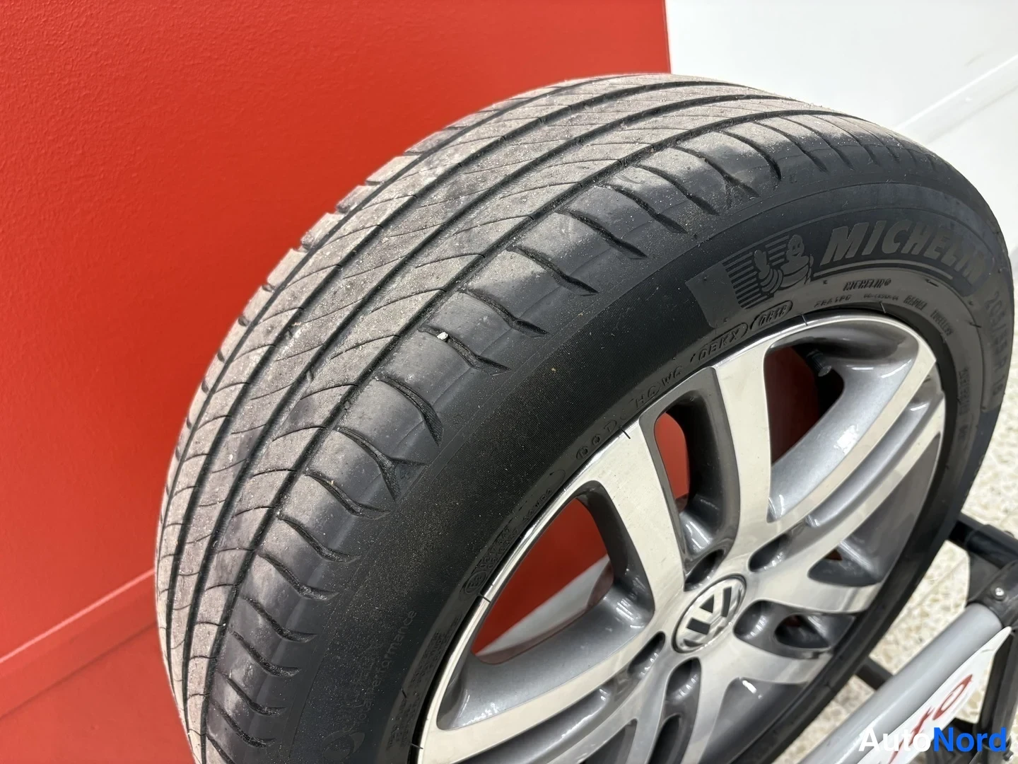 Yksityiskohta: lähikuva auton renkaasta ja hopeanvärisestä alumiinivanteesta; renkaan kyljessä lukee Michelin, ja vanteessa on Volkswagen-merkki. Taustalla punainen seinä.