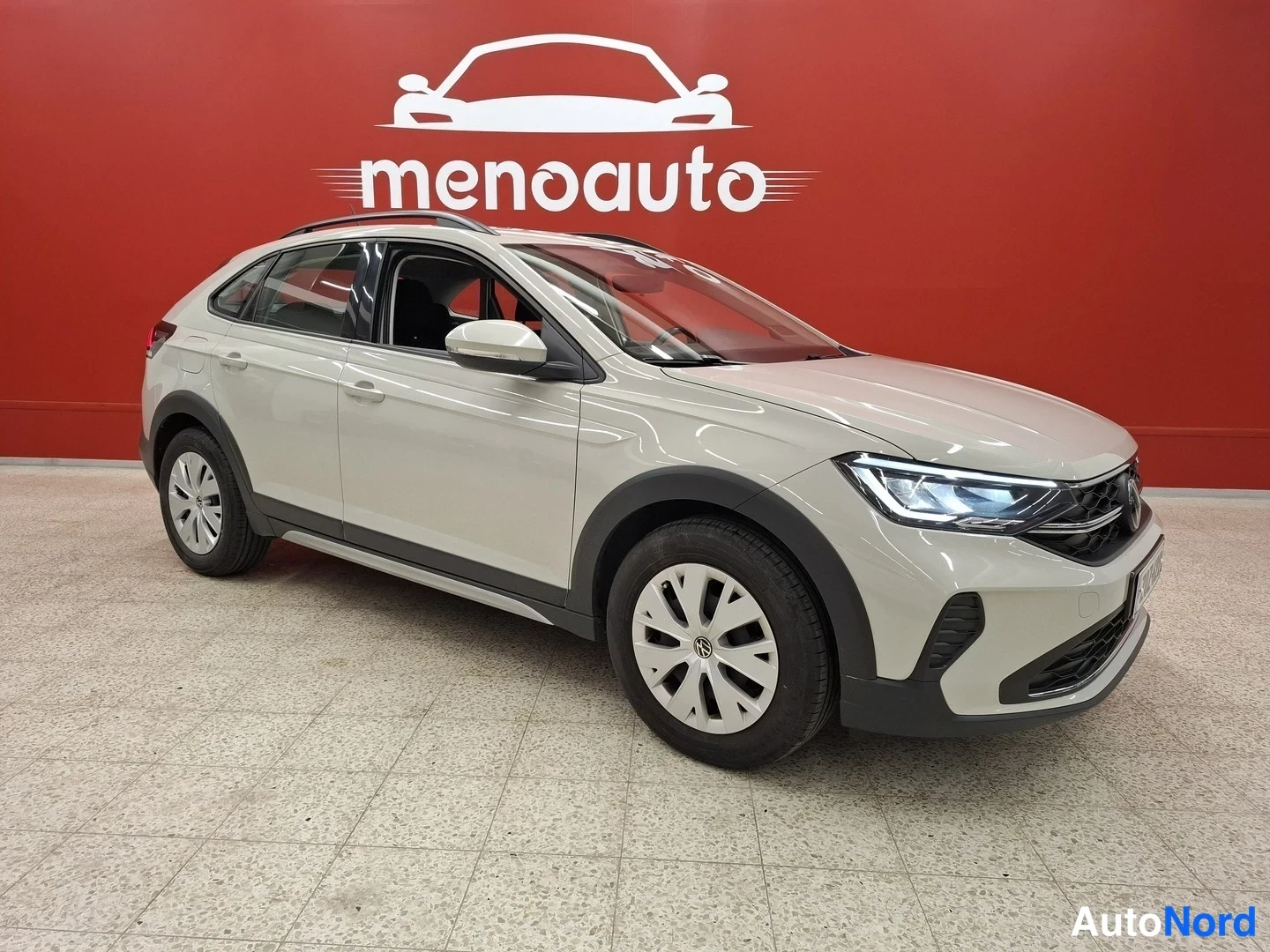 Etuviisto, nokka oikealle: Vaaleanbeige Volkswagen Taigo -henkilöauto kuvattu etukulmasta oikealta punaisen seinän edessä.