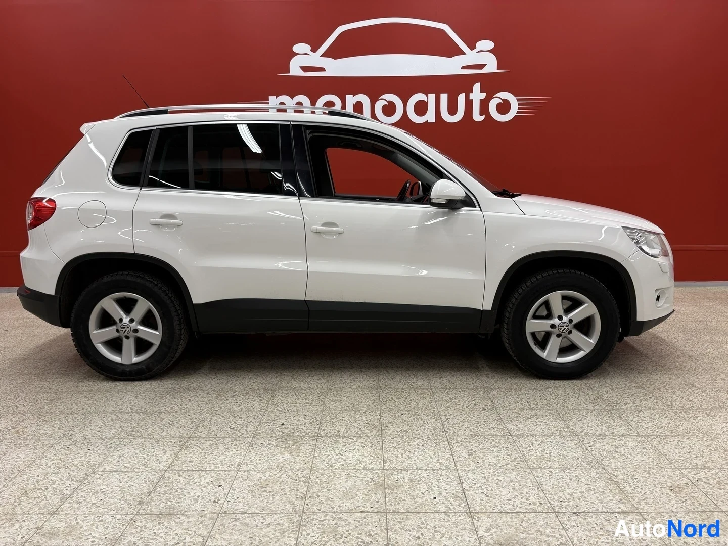 Etuviisto, nokka oikealle: Valkoinen Volkswagen Tiguan sivulta oikealta nähtynä, punainen menoauto-taustaseinä.