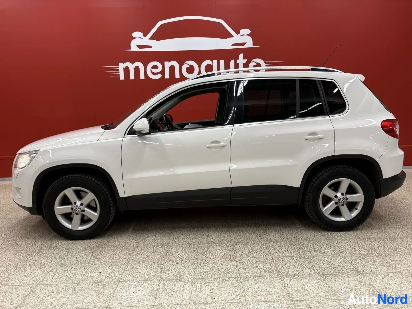 Etuviisto, nokka vasemmalle: Valkoinen Volkswagen Tiguan sivulta etukulmasta sisätiloissa, kylki ja keulanosa näkyvät.