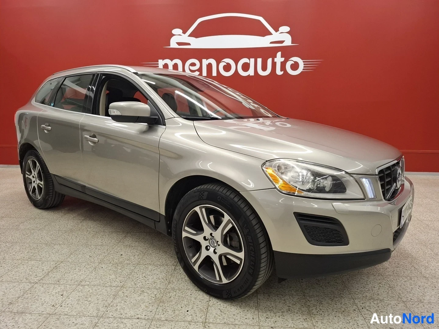 Etuviisto, nokka oikealle: Volvo XC60 -farmari kuvattu kolmannelta etukulmalta, keula oikealla, kylki vasemmalla, messiinharmaa koria punaisella taustalla.