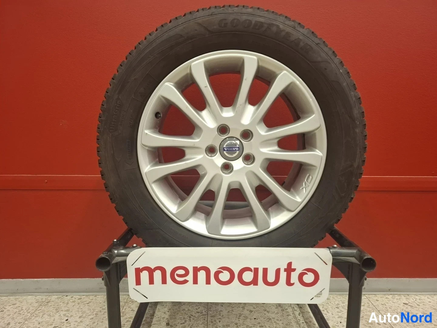 Yksityiskohta: Hopeanvärinen Volvo-alkuvuu tilalla punaisella seinällä taustalla; rengas on Goodyear talvirengas.