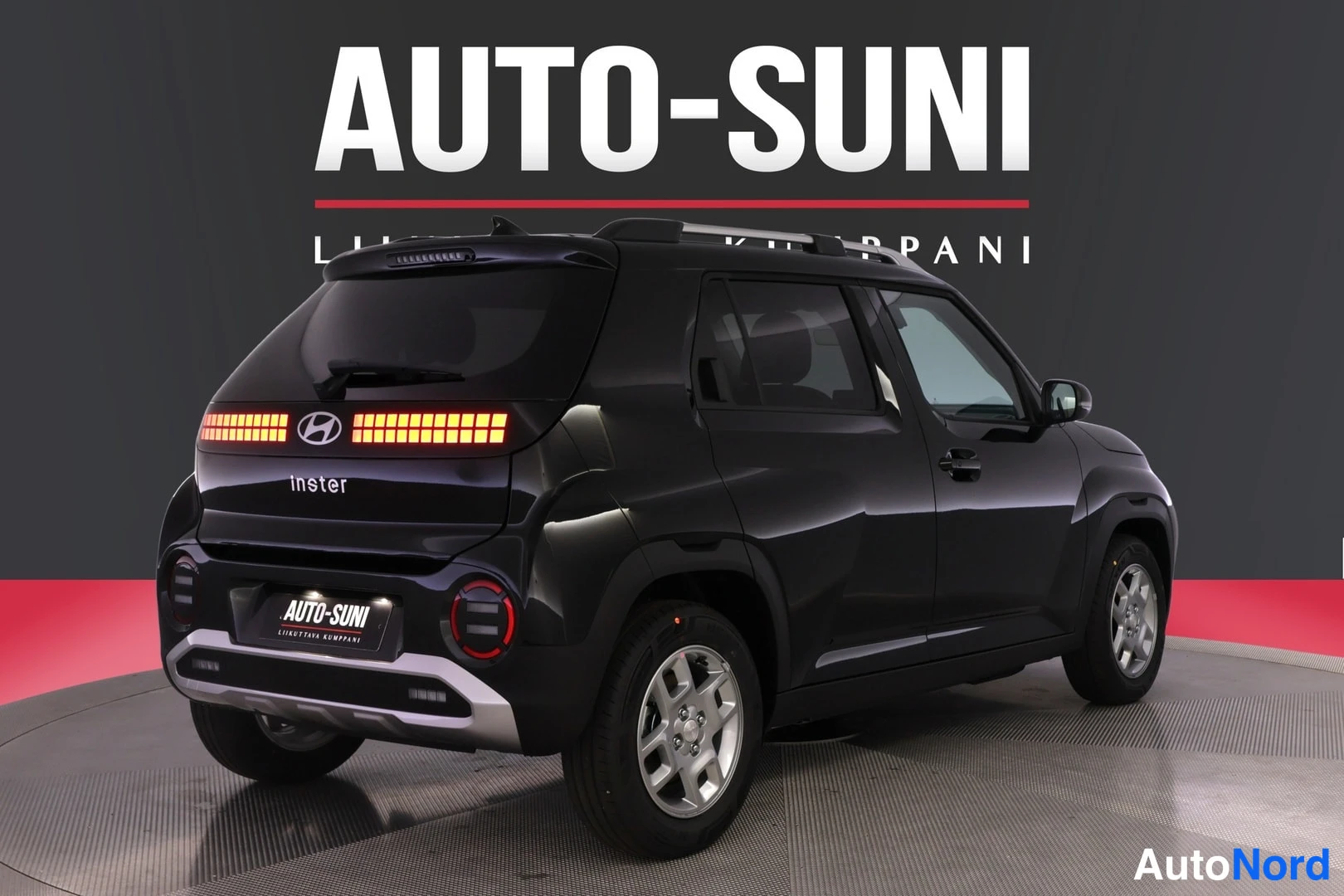 Takaviisto: Takakolmannes vasemmalta mustasta autosta; pinnan ja lasin takana näkyy Inster-teksti, takavalot, Hyundai-logo, AUTO-SUNI-kyltti.
