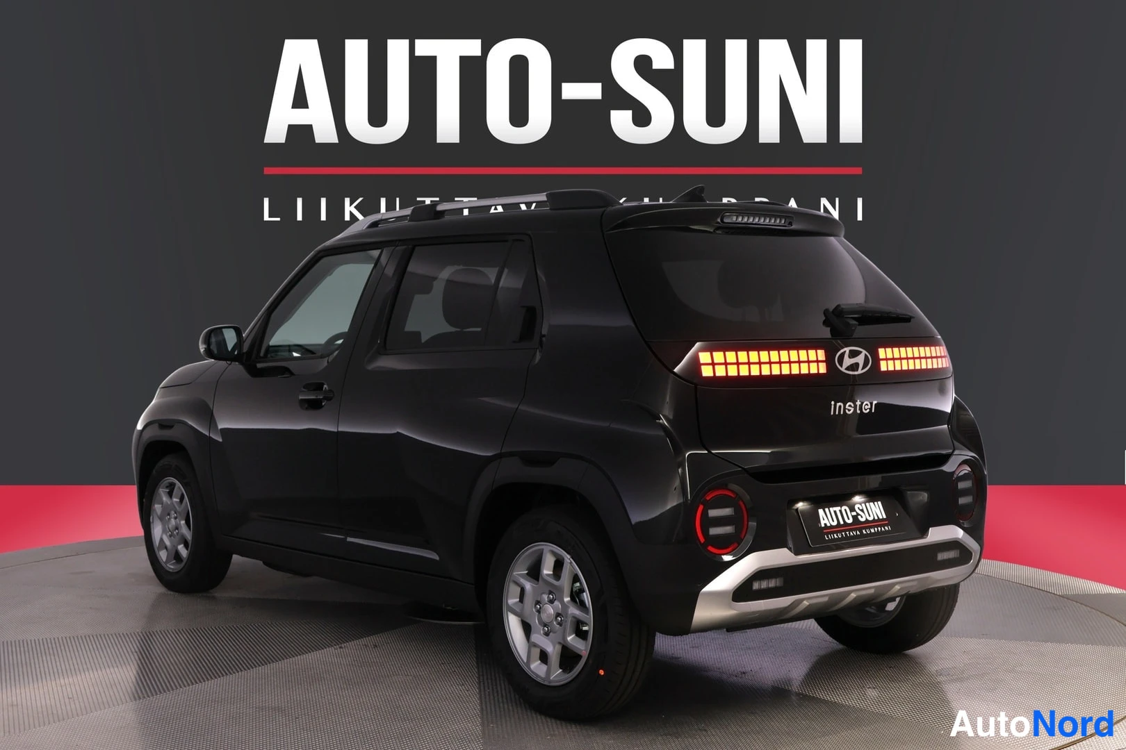 Takaviisto: Takakolmannes oikealta mustasta autosta; takavalot ja Hyundai-logo, takana AUTO-SUNI-kyltti, kuljettajan puoli ja alumiinivanteet näkyvät.