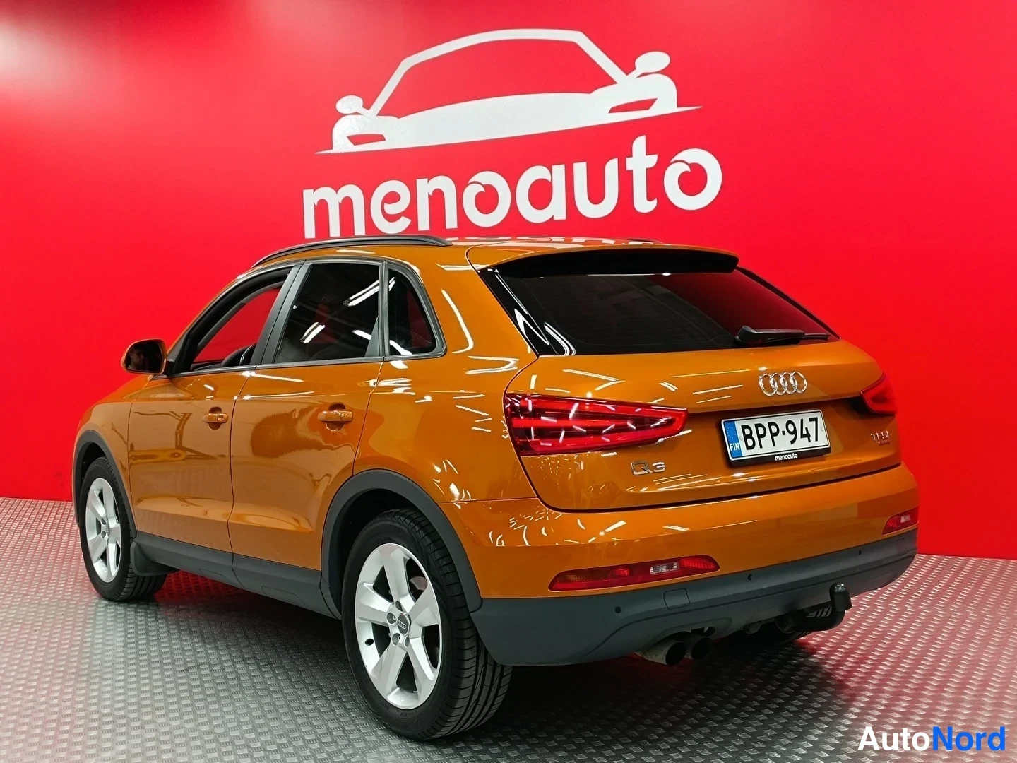 Takaviisto: Oranssi Audi Q3 -henkilöauto kuvattu taka kolmasosakulmasta autonokki oikealla, punainen menoauto-taustaseinä.