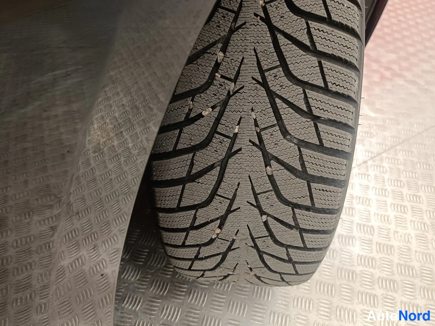 Yksityiskohta: Lähikuva auton renkaasta: talvirengas urillaan, jonka urissa on kivimurskaa. Auto seisoo ristipeltialustalla.