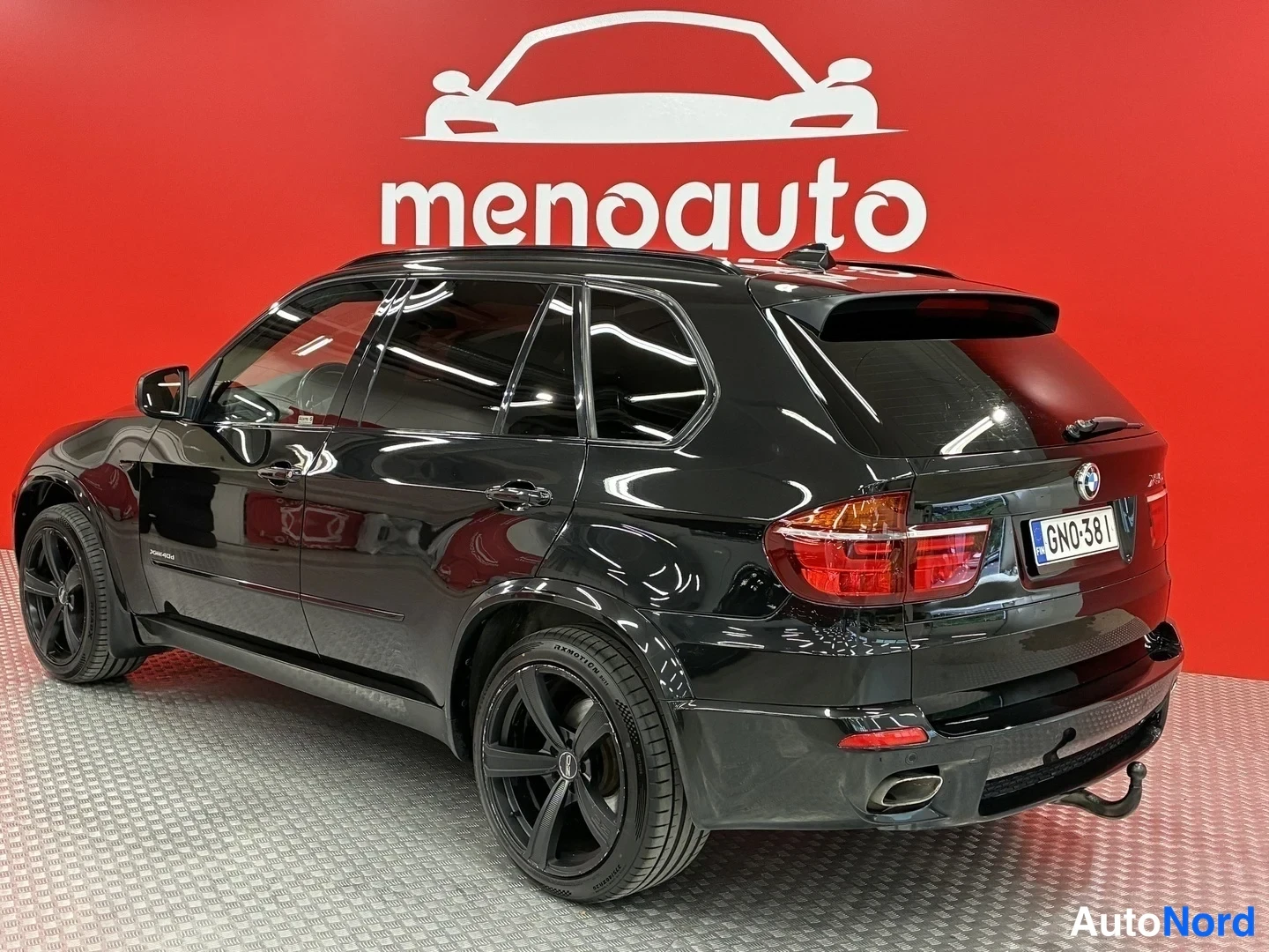 Takaviisto: BMW X5:n takakulmanäkymä: musta autoc näkyy takavaloineen, takapuskineen ja vasemman puolen ovineen punaisella taustaseinällä.