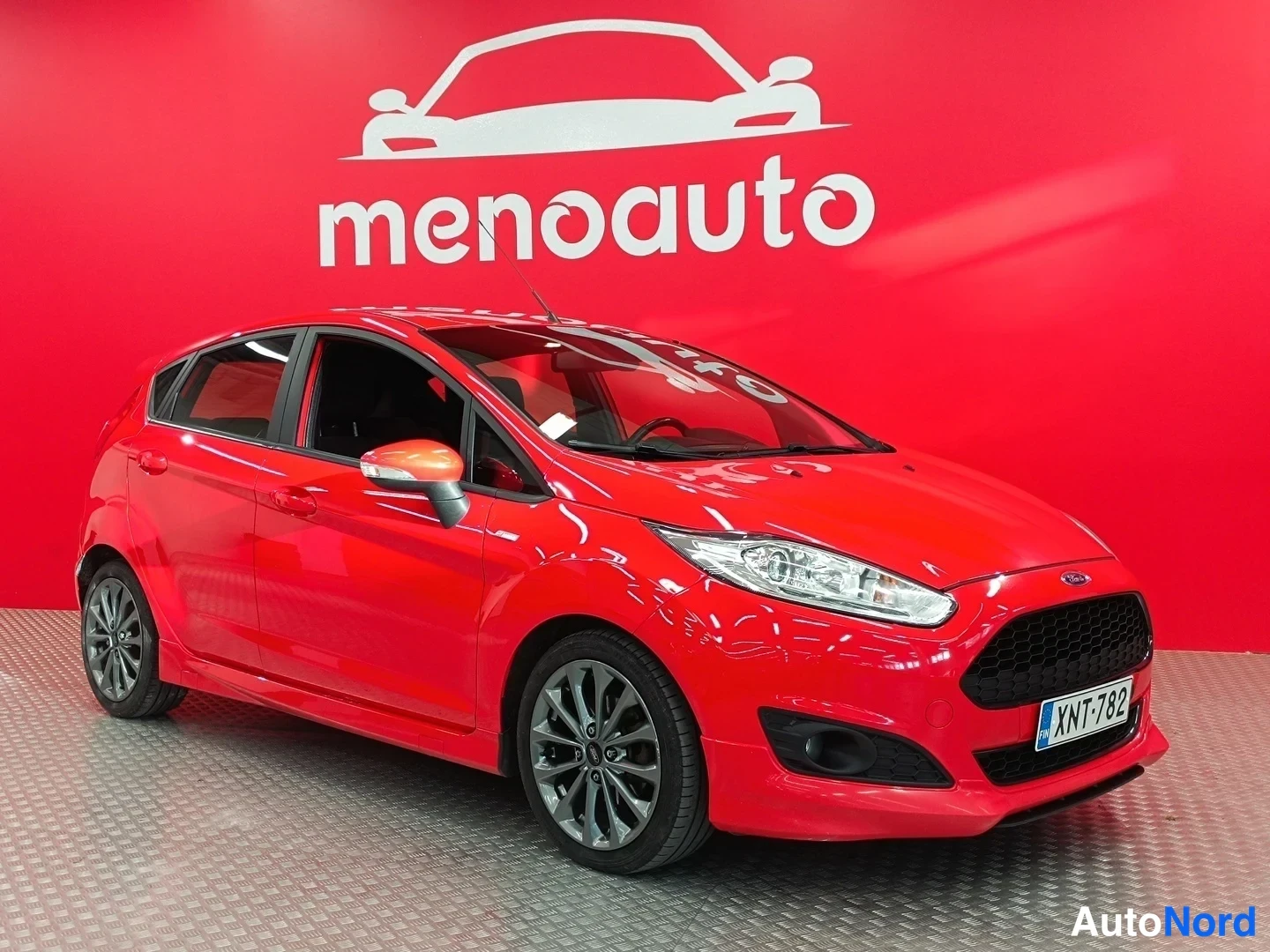 Etuviisto, nokka vasemmalle: Punainen Ford Fiesta etuviistosta oikealta, rekisteritunnus, valokuvattuna sisätiloissa punaisella menoauto-taustaa vasten.