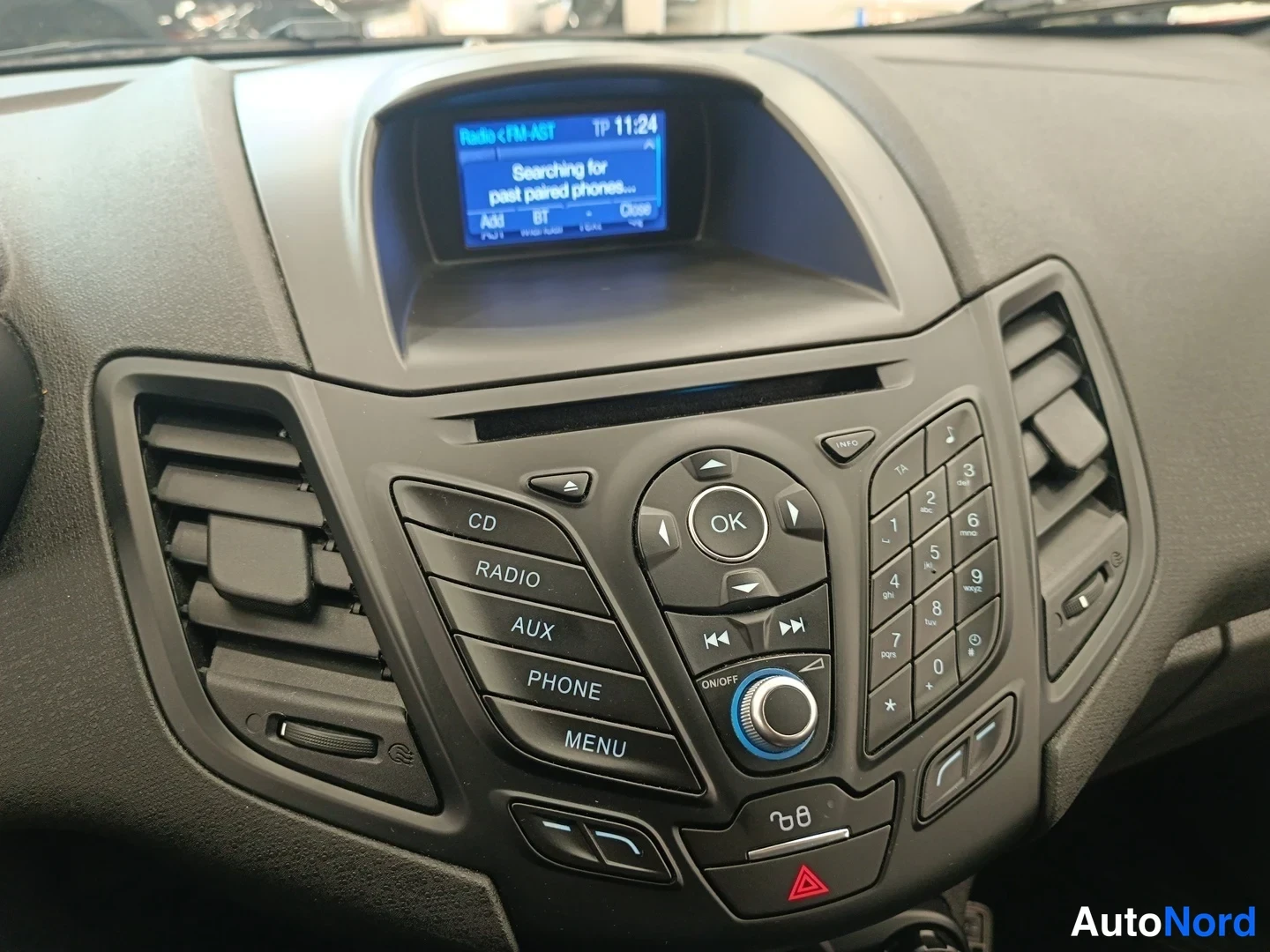 Muu: Lähikuva auton keskikonsolin radiosta ja näytöstä, jossa näkyy Radio FM-ABT, TP 11:24 ja teksti 'Searching for past paired phones...'. Napit CD, RADIO, AUX, PHO