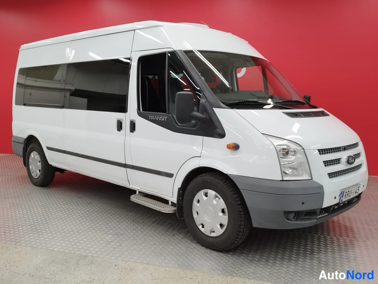 Etuviisto, nokka oikealle: Valkoinen Ford Transit -pakettiauto kolmesta neljäsosasta edestä oikealta, korostettu etusäleikkö ja sivuovi, punainen tausta.