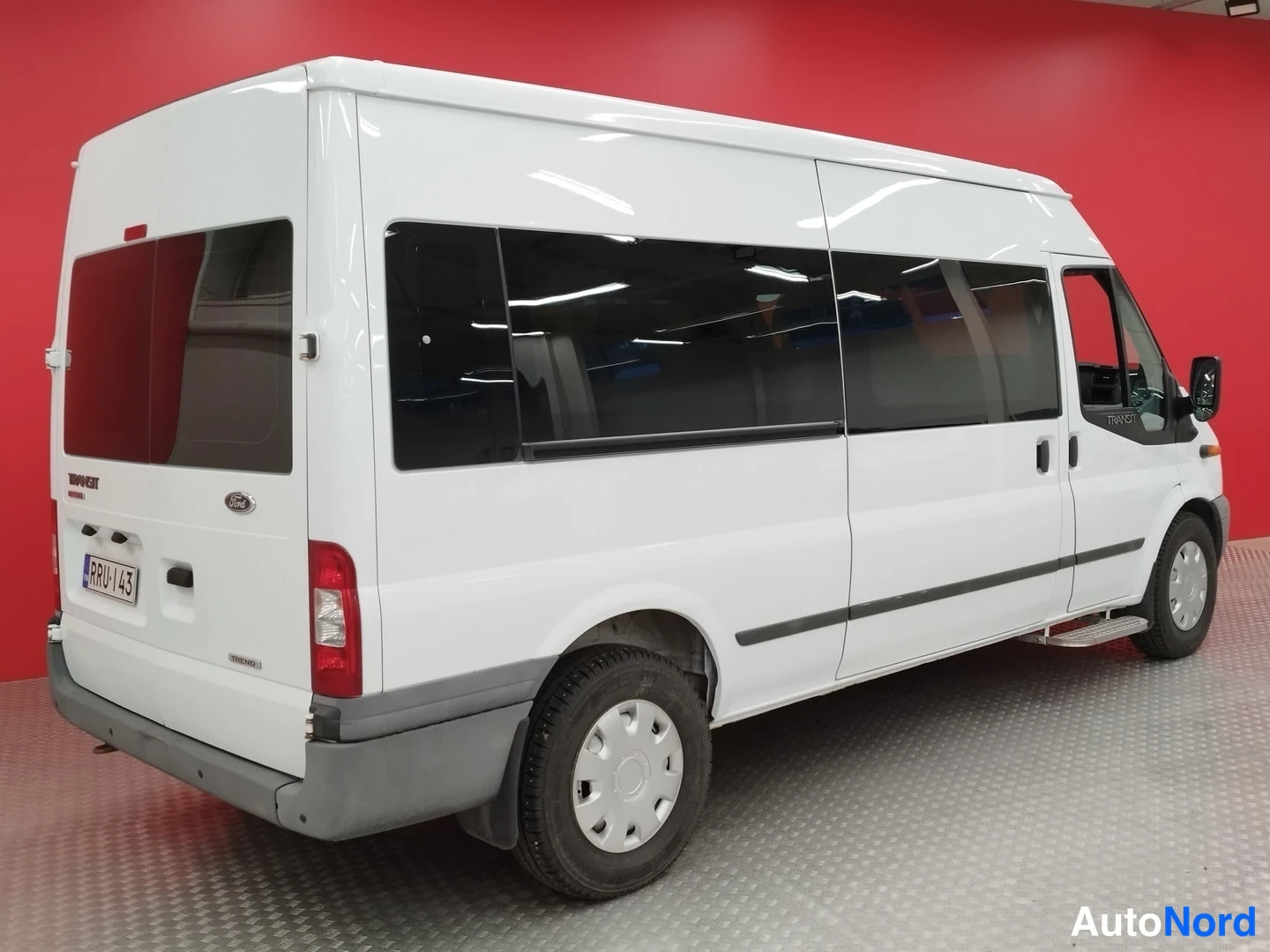 Takaviisto: Valkoisen Ford Transit -pakettiauton takaosa ja vasen kylki takaa vasemmalle kuvattuna, tummat ikkunat ja rekisterikilpi, punainen tausta.