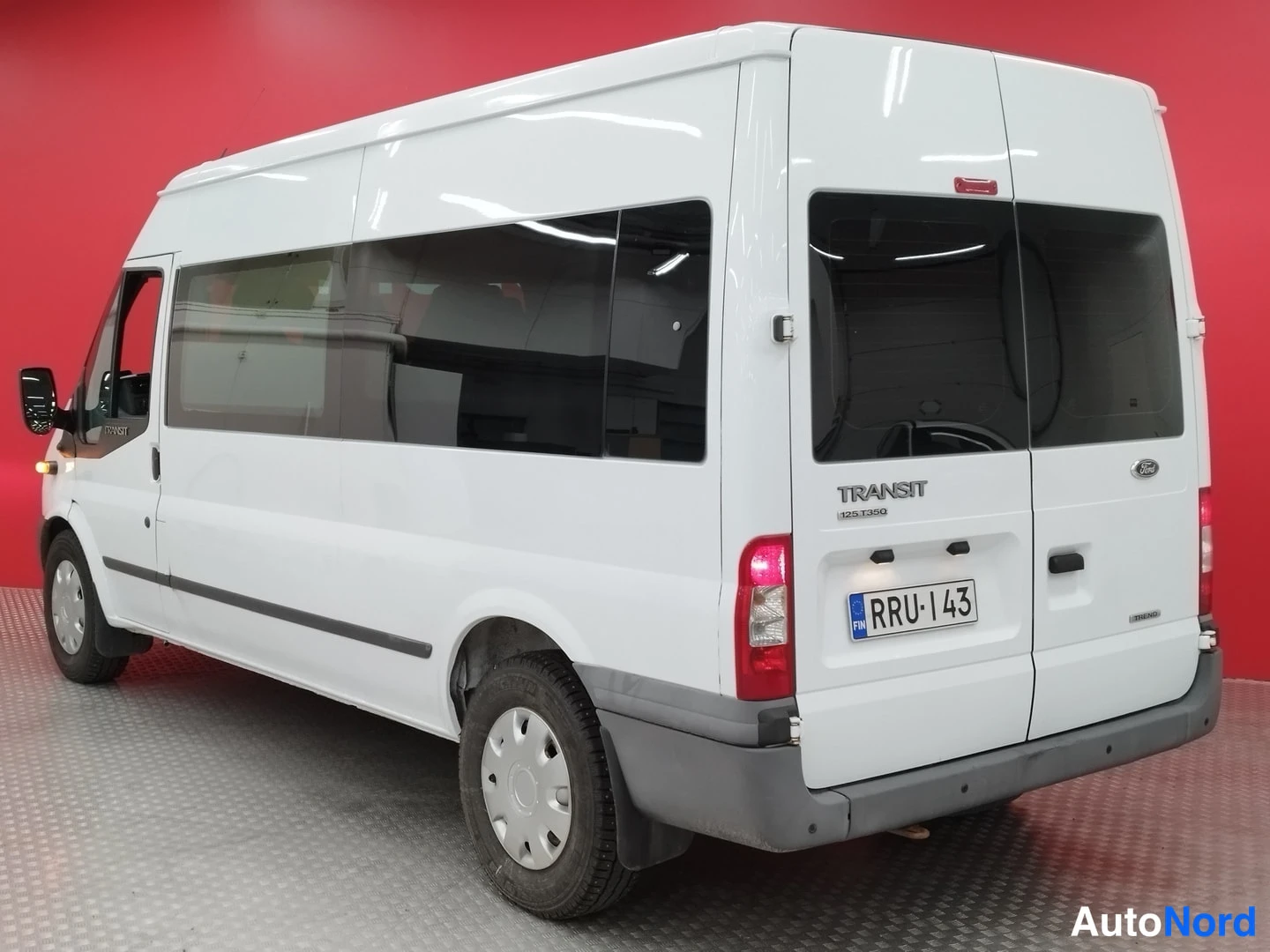 Takaviisto: Valkoisen Ford Transit -pakettiauton takaosa ja vasen kylki takaa vasemmalle kuvattuna, TRANSIT-teksti ja rekisterikilpi takaluukussa, punainen tausta.