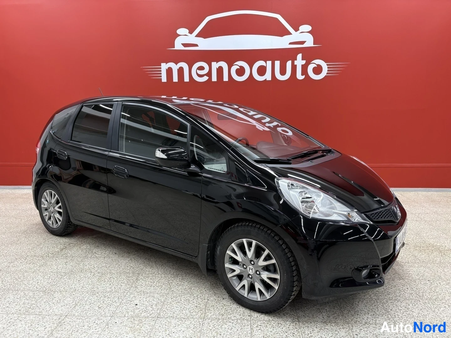 Etuviisto, nokka vasemmalle: Musta Honda Jazz viistosti edestä oikealta kuvattuna sisätiloissa punaisella taustaseinällä.
