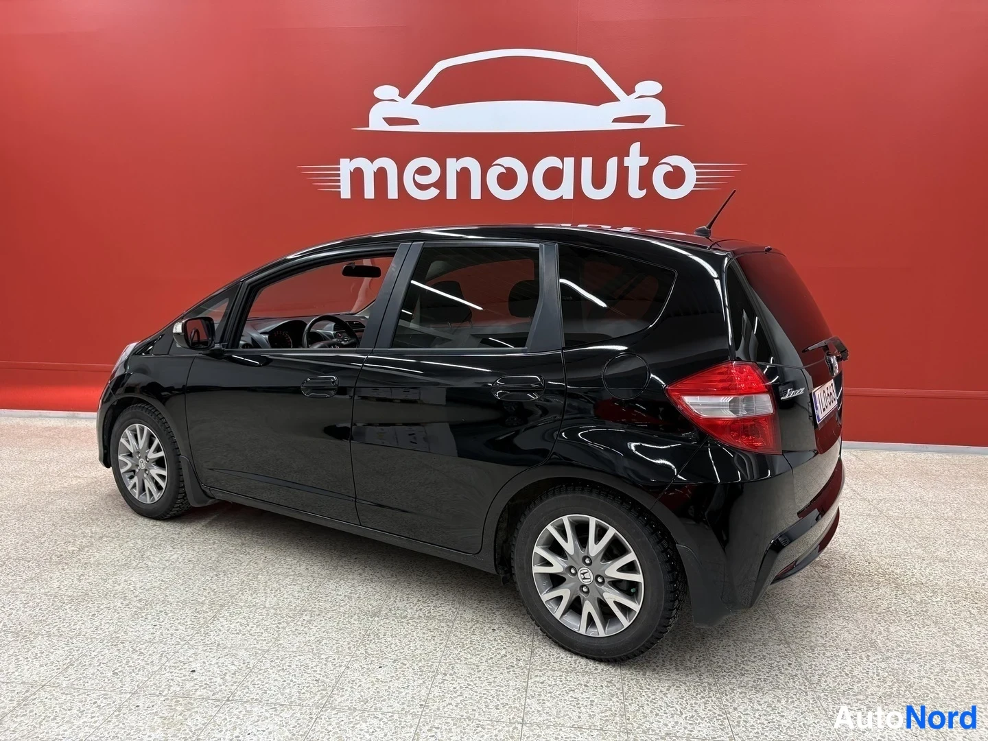Takaviisto: Mustan Honda Jazzin takakolmannesvasen näkymä sisätiloissa, punainen menoauto-taustaseinä ja vaalea laattalattia.