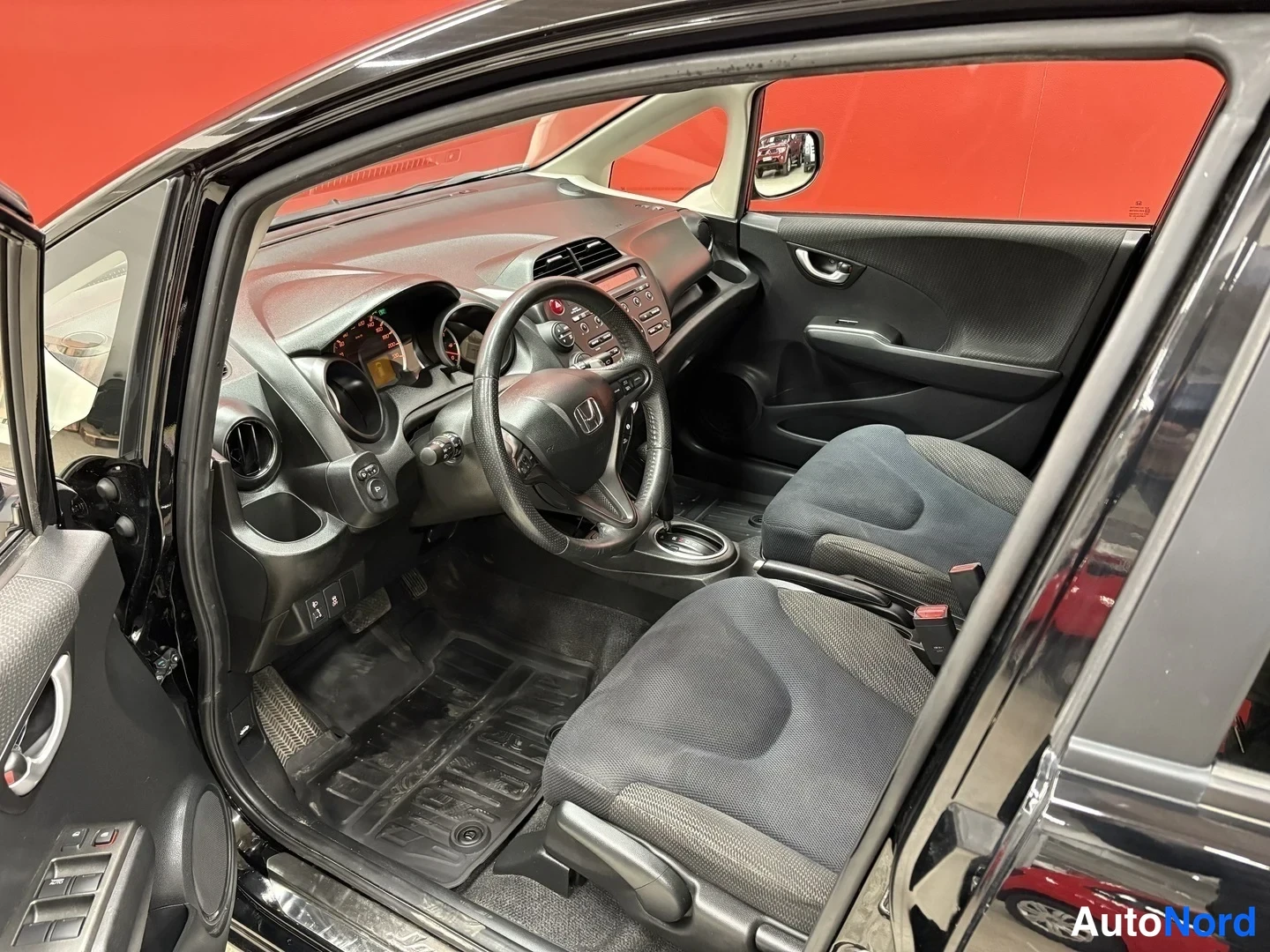 Muu: Honda Jazzin etuistuimet ja kojelauta kuljettajan ovi auki kuvattuna; tummaa kangasverhoilua ja Honda-ohjauspyörä.