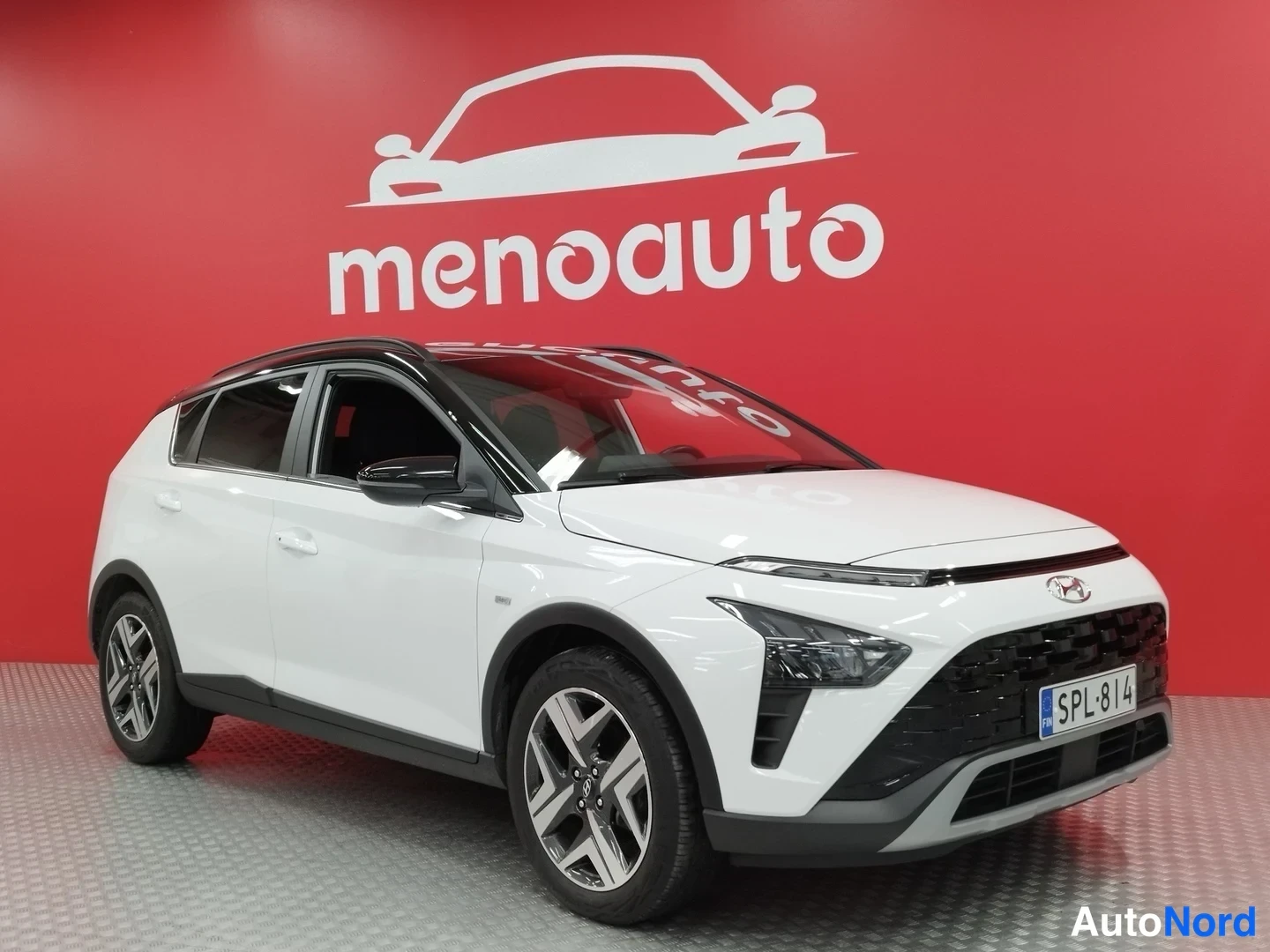 Etuviisto, nokka oikealle: Valkoinen Hyundai Bayon kulmassa edestä oikealta, musta katto ja konepellin LEDs-sarvi, rekisteri, punainen studioseinä taustalla.