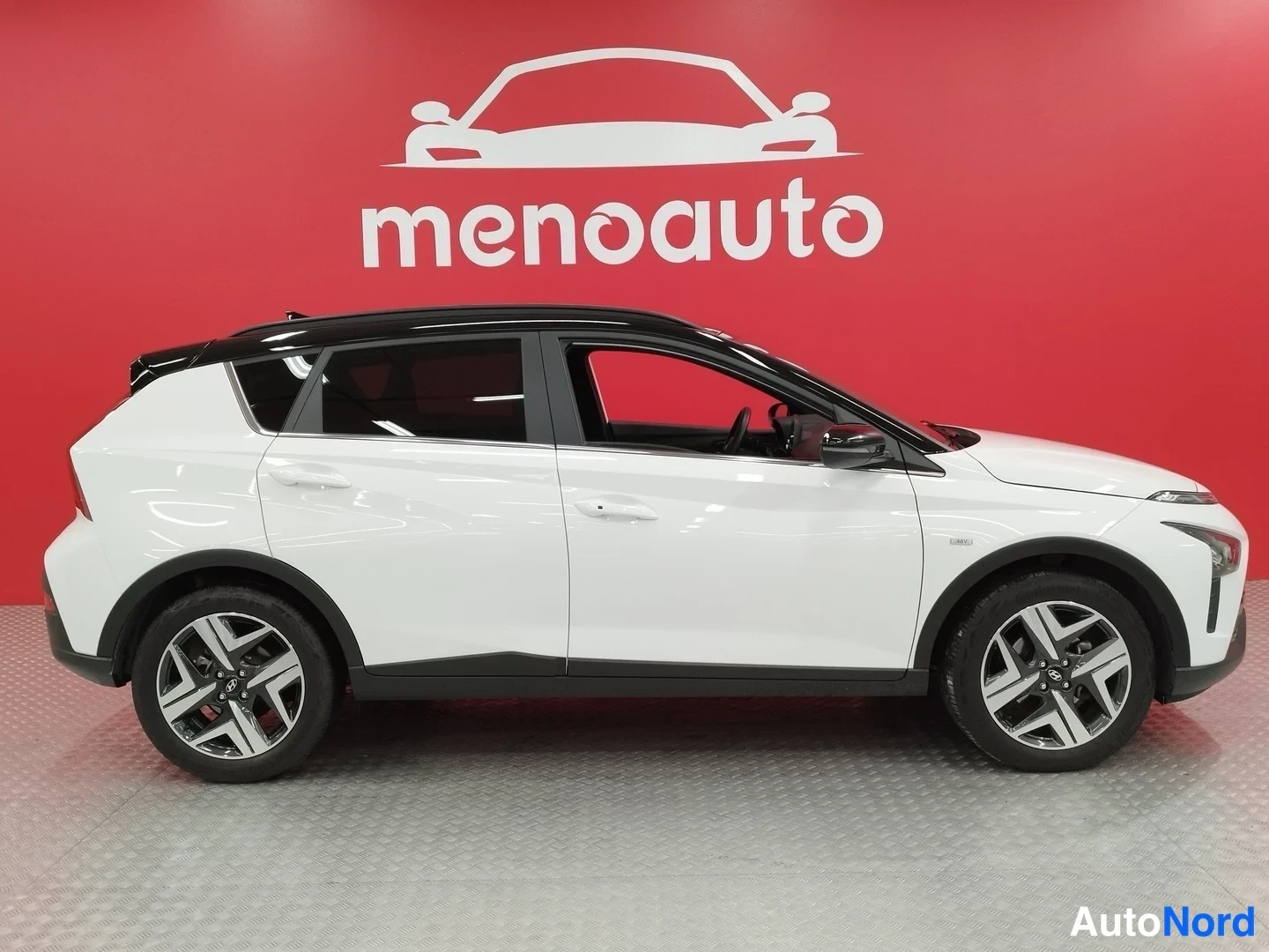 Etuviisto, nokka oikealle: Valkoinen Hyundai Bayon sivulta oikealta, musta katto ja viisiovainen korit, punainen menoauto-tausta.