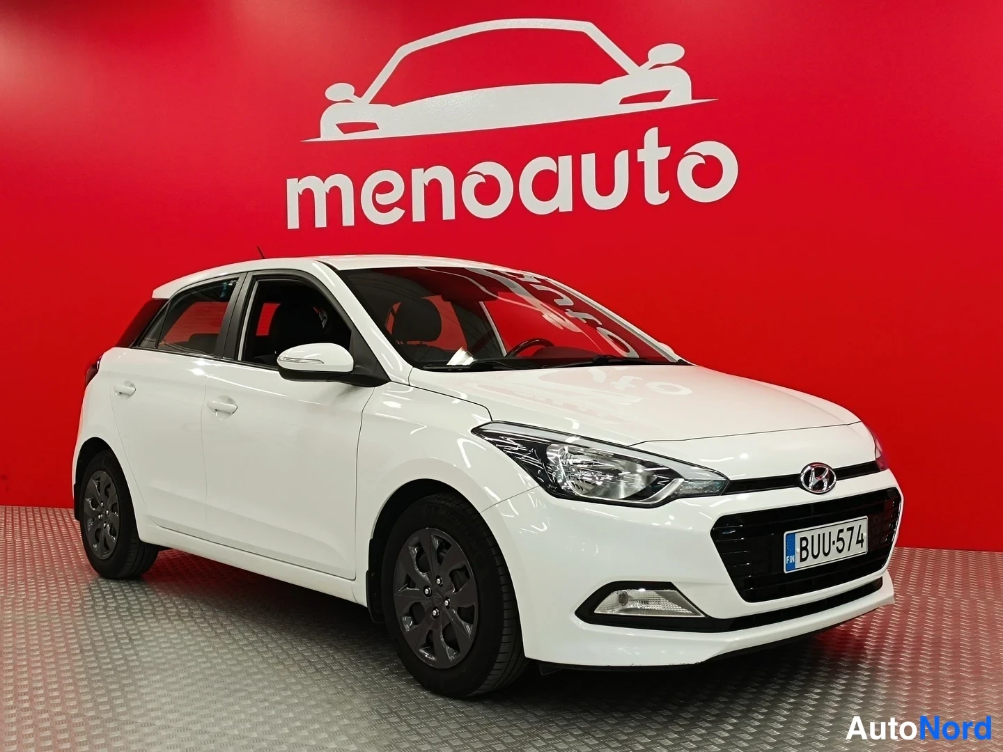 Etuviisto, nokka oikealle: Valkoinen Hyundai i20 -viistotakaa edestä oikealta, rekisteri, punainen menoauto-tausta.