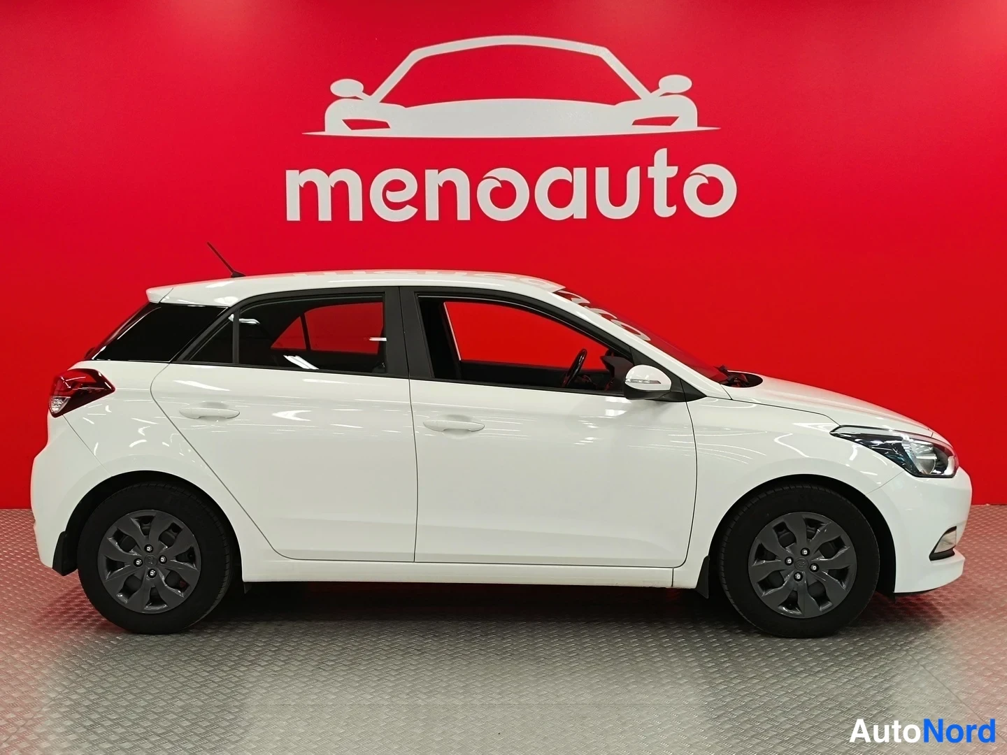 Sivulta: Valkoisen Hyundai i20 -henkilöauton kylj edestä oikealta, punainen menoauto-tausta.