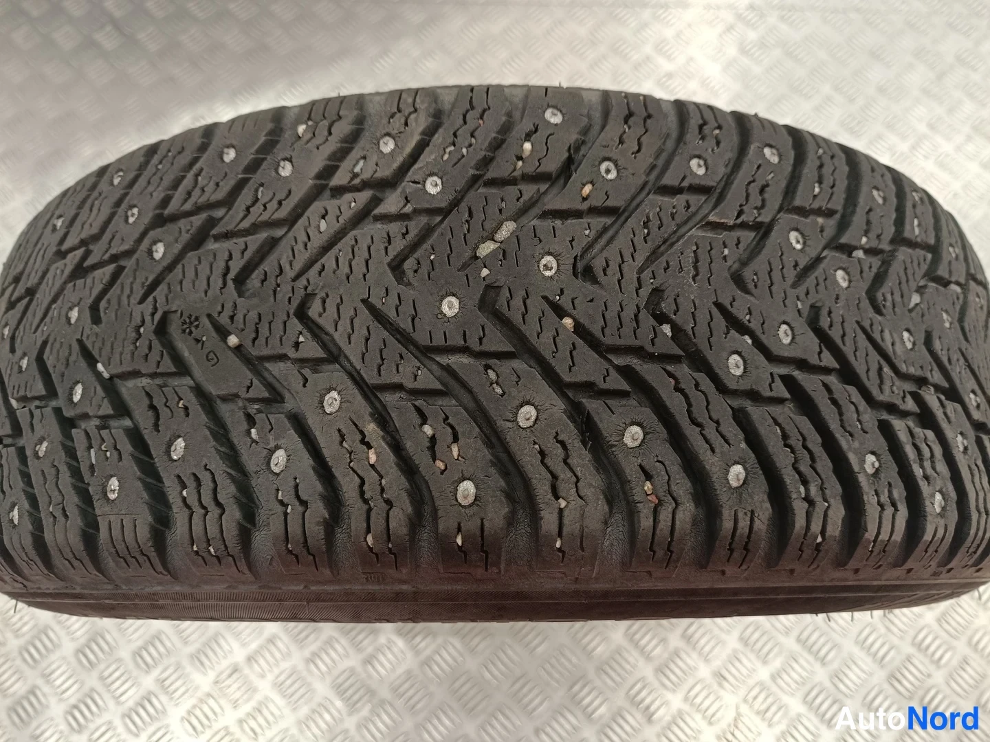Sivulta: Closeup of a black studded winter tyre: the tread pattern with ice-boost sipes and studs is visible against a patterned aluminium background.