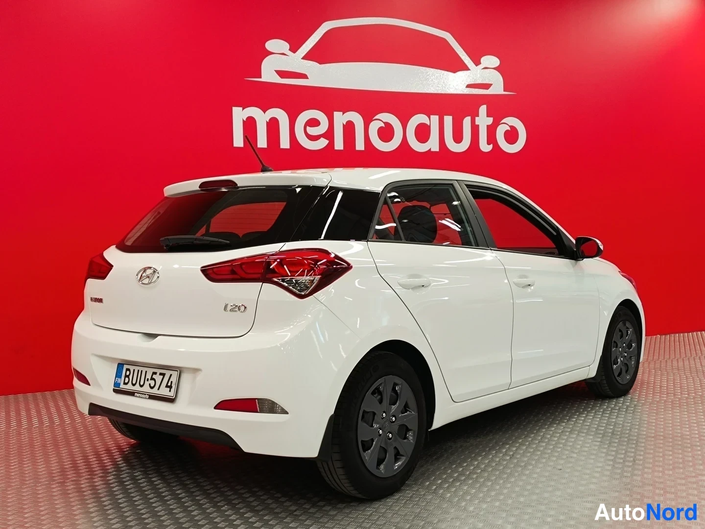 Takaviisto: Valkoisen Hyundai i20 -auton takaosa ja kylki viistosti takaa vasemmalta, rekisteri, punainen menoauto-tausta.