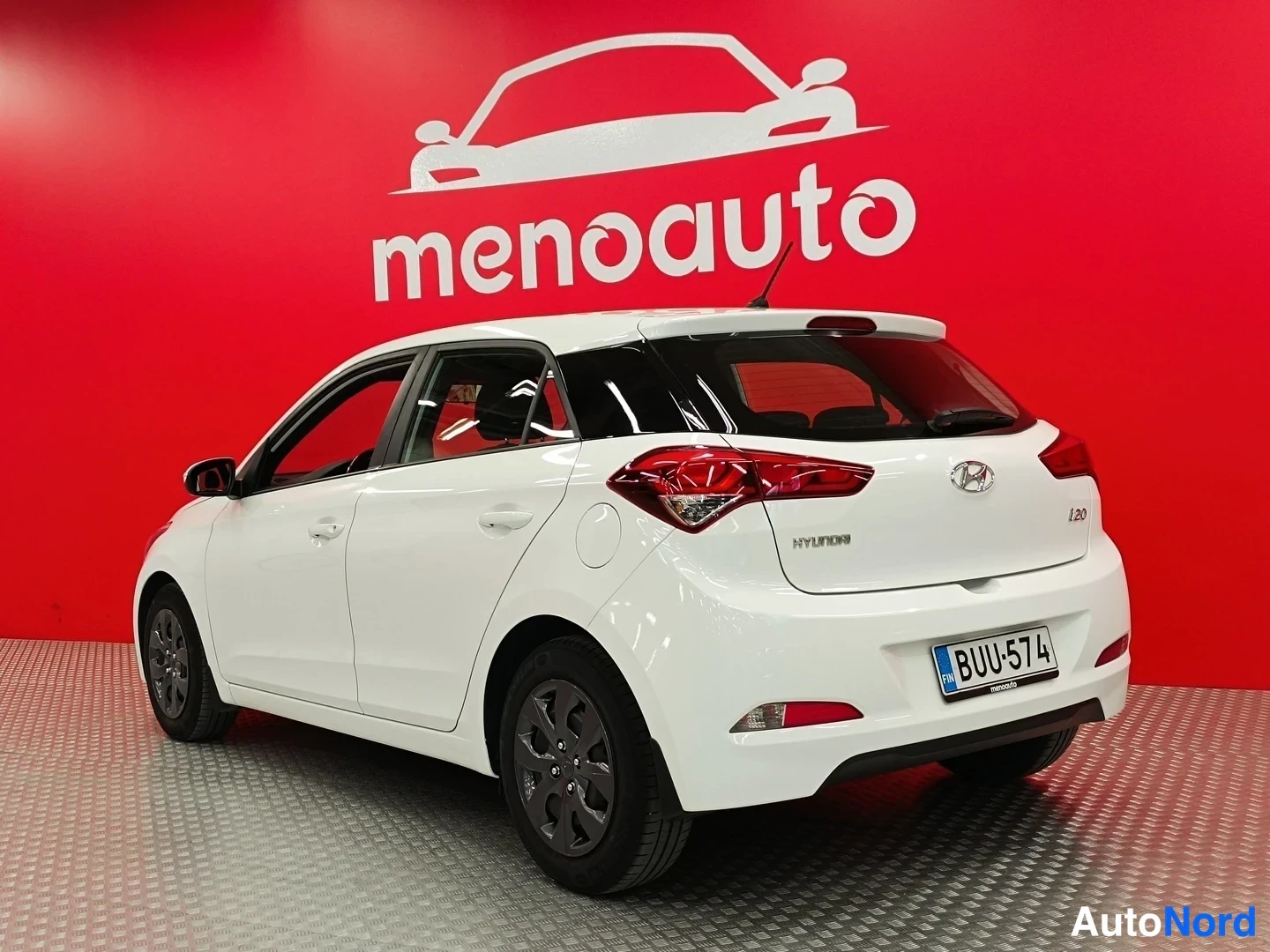 Takaviisto: Valkoinen Hyundai i20 takaviistosta, rekisterikilpi, punainen menoauto-taustaseinä.