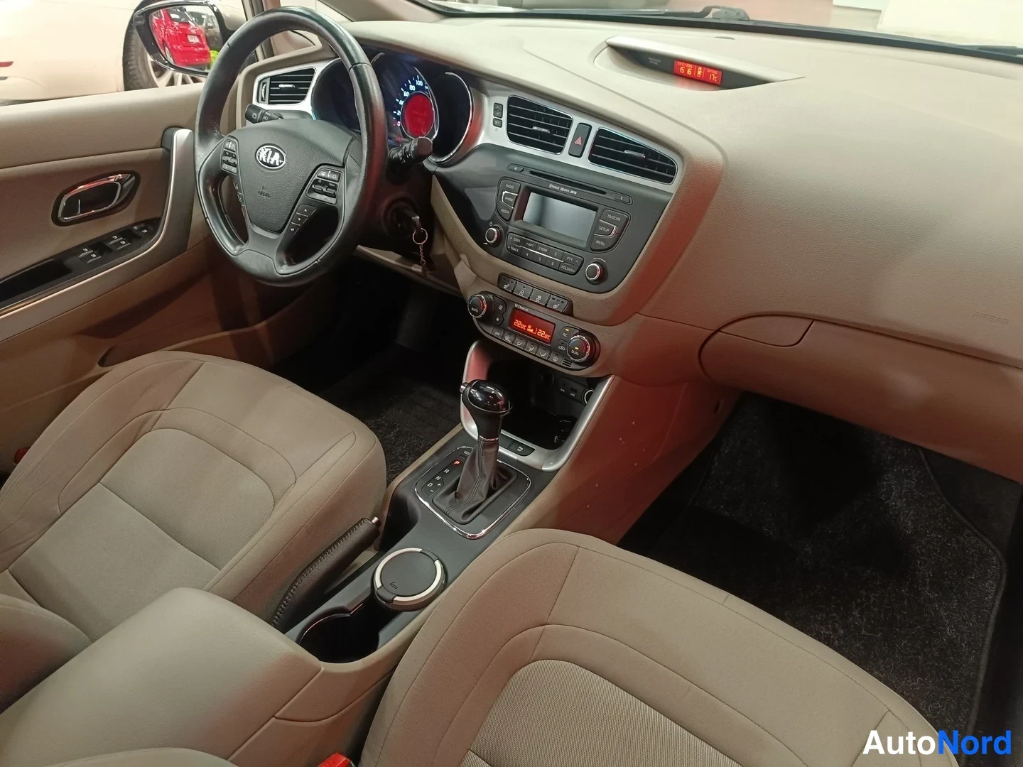 Muu: Auton etuistuimilta kuvattu kojelauta: beige(panel)dashboard, KIA-ohjauspyörä, äänentoistojärjestelmän paneeli, automaattivaihteen valitsin ja kellonäytössä aik