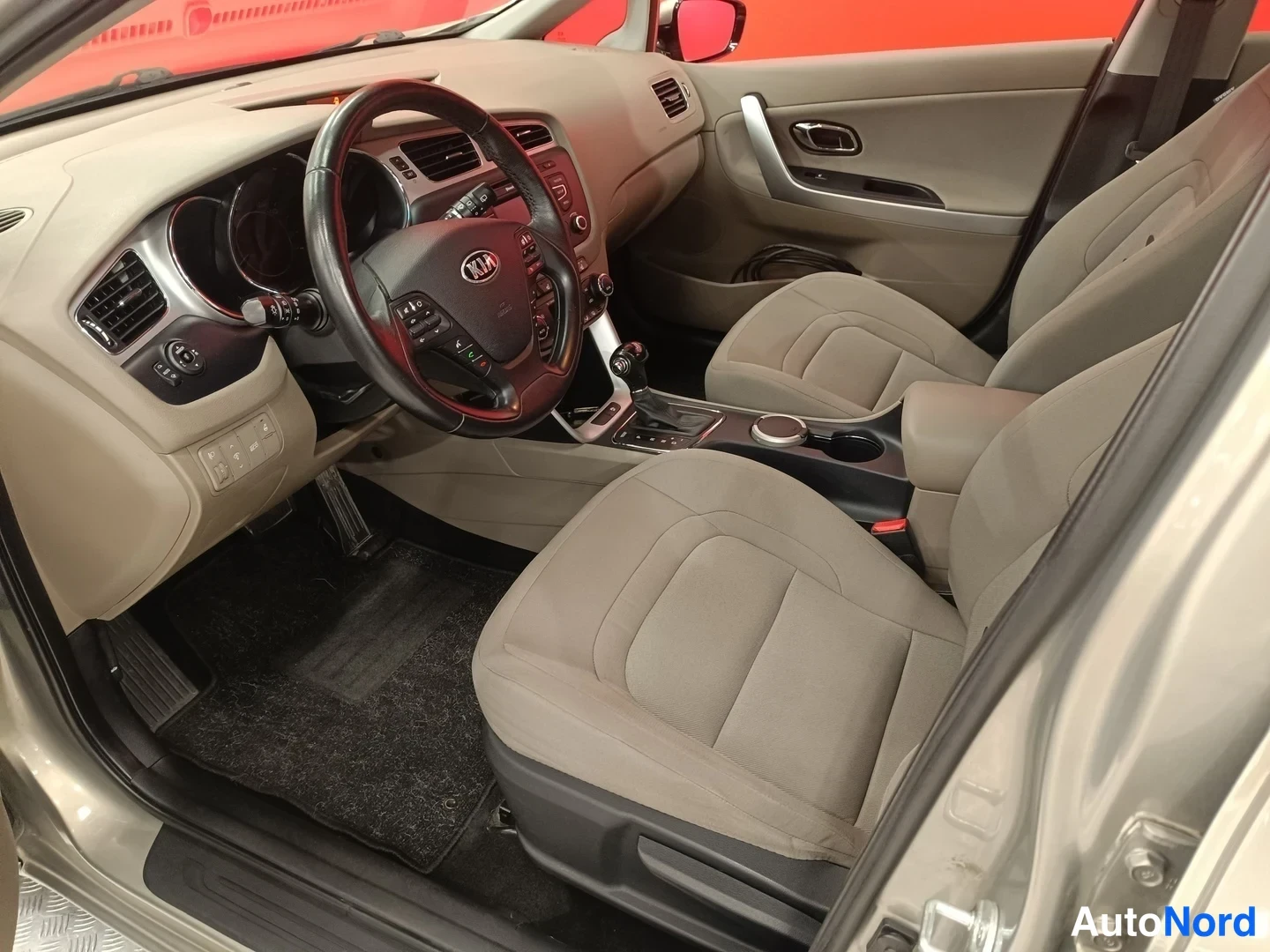 Muu: Kia-merkkisen auton sisätilat kuljettajan puolelta: beige konepelti-istuimet, monitoimiohjauspyörä, automaattivaihteen valitsin ja KIA-logo ohjauspyörässä.