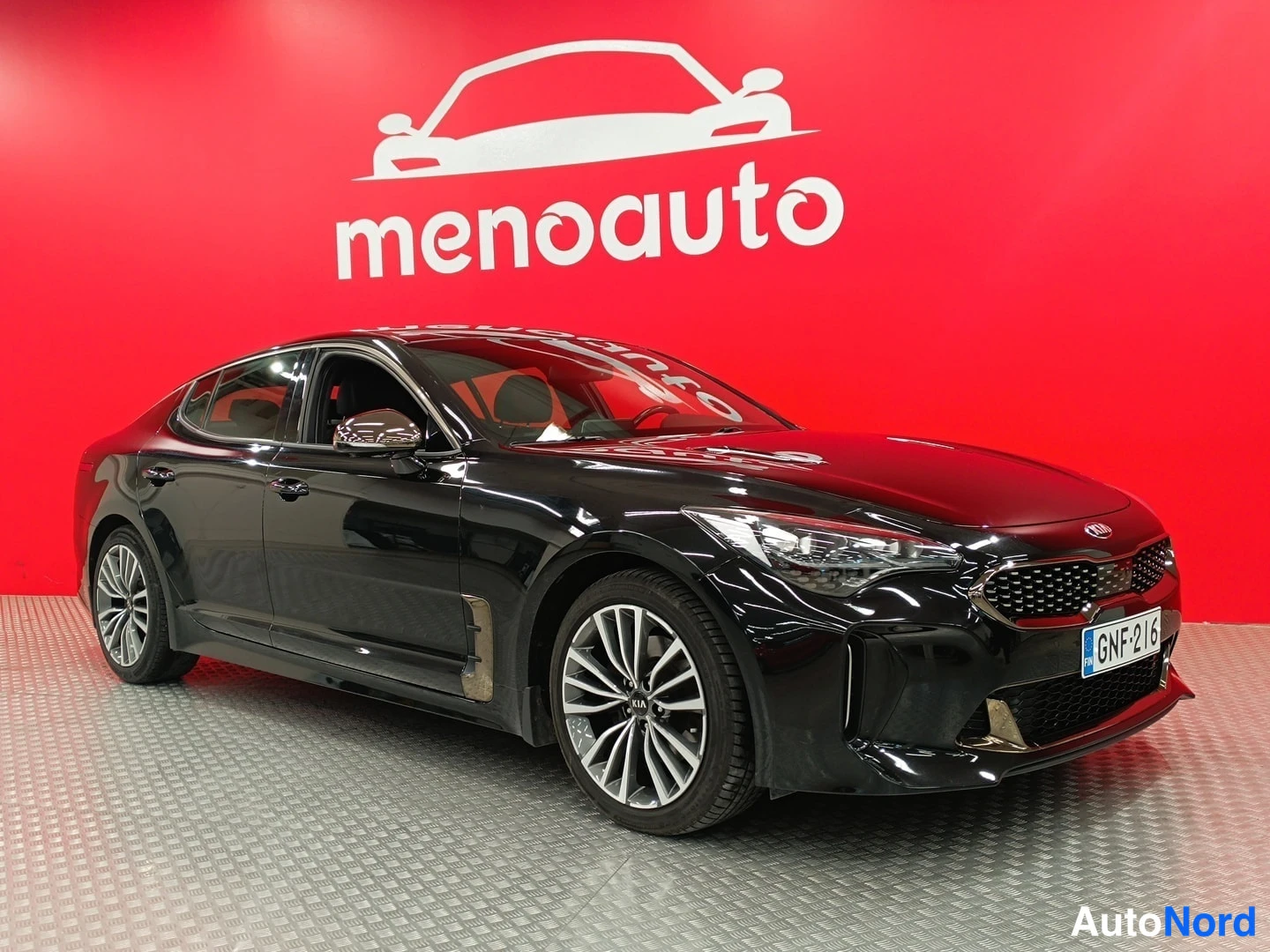 Etuviisto, nokka oikealle: Mustan Kia Stinger -farmarin(?) etukulma oikealta, punainen tausta